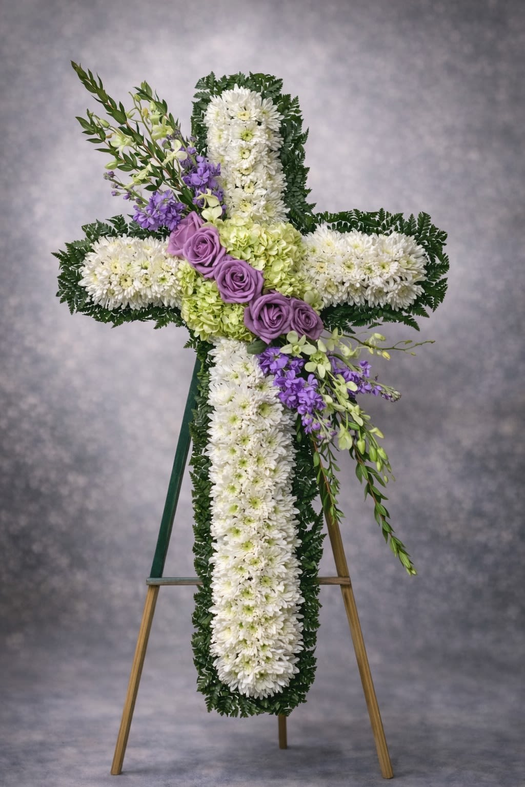 Garden of Peace Cross - A graceful standing cross designed with fresh white chrysanthemums, soft lavender roses, hydrangea accents, and delicate seasonal blooms. This elegant tribute symbolizes peace, faith, and everlasting love.  Displayed on an easel, this arrangement offers a comforting and serene presence at funeral services, memorials, and church ceremonies.  Substitutions may be necessary based on seasonal availability, but the overall cross shape, color harmony, and fullness will always be maintained.  Delivery available to funeral homes, churches, cemeteries, and residences. Please include service time and location details at checkout.