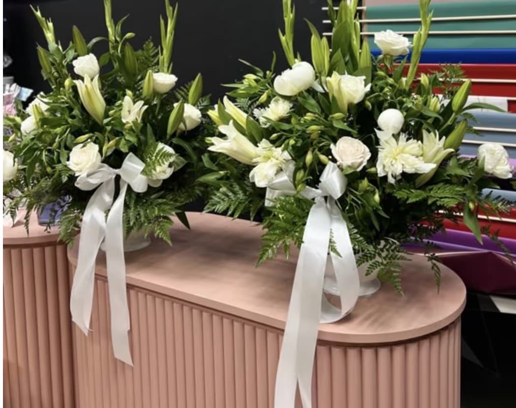 Custom orders  - Two matching white floral arrangements that read as serene and refined-perfect as a sympathy tribute, wedding accent, or a calming everyday gift. These formal bouquets combine Asiatic lilies, white roses, chrysanthemums and other daisy-like blooms arranged in a full, rounded shape so the person you're thinking of will feel comforted and honored. The white colorway and polished presentation convey quiet elegance for moments that need gentle presence.  Each arrangement is finished with abundant green foliage, a long white ribbon, and set in a clean white container for a cohesive, ceremonial look. The compact, rounded design makes these easy to place on a mantel, reception table, or beside a bedside-designed to feel composed from every angle. Order as a matching pair for a balanced display; these arrangements arrive ready to present with minimal care needed-keep fresh water in the containers and trim stems if repotted to extend bloom life.