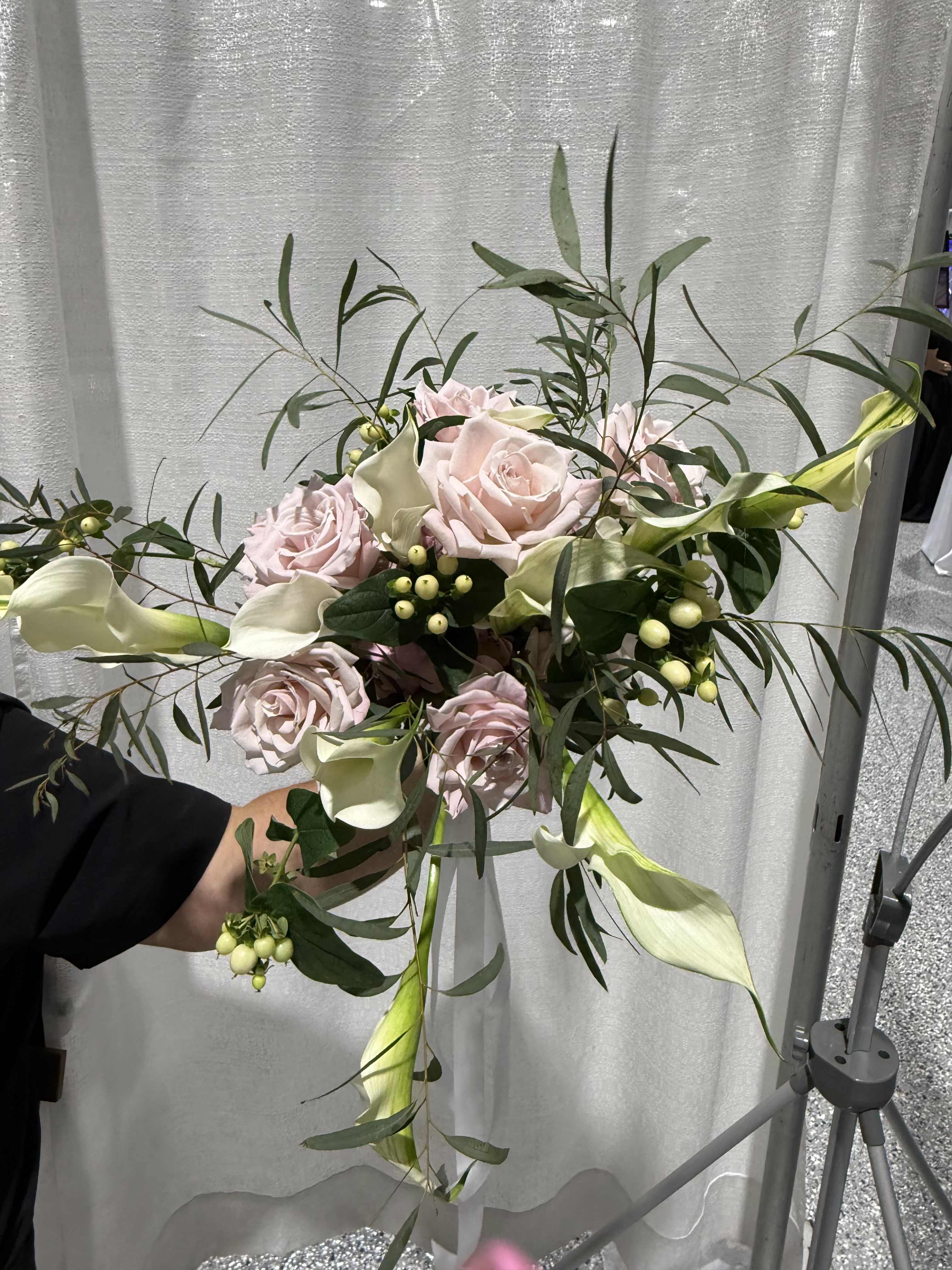 Soft blush  - Soft blush roses and sculptural white calla lilies come together in an airy, asymmetrical hand-tied bouquet that feels like a modern garden picked at the peak of bloom. This wedding bouquet and romantic gift option reads elegant and refined-the pale pink and ivory palette, with long slender foliage and small pale berry clusters, creates a delicate, heartfelt impression that will make the person you're thinking of feel truly seen.  The arrangement pairs velvety rose heads with the smooth, trumpet shapes of calla lilies, accented by elongated greenery and subtle berry sprigs for texture and movement. The loose, hand-tied design gives it a natural silhouette that drapes slightly rather than sitting rigidly, ideal for brides seeking effortless romance or for marking a special anniversary or celebration with quiet sophistication.  Perfect for weddings, anniversaries, or a thoughtful romantic surprise, this bouquet arrives ready to place in a vase or carry down the aisle; minimal care-fresh water and a cool spot-keeps blooms looking their best for several days. If you want to ensure the most flattering fit for the occasion, select your preferred size at checkout for the right scale.