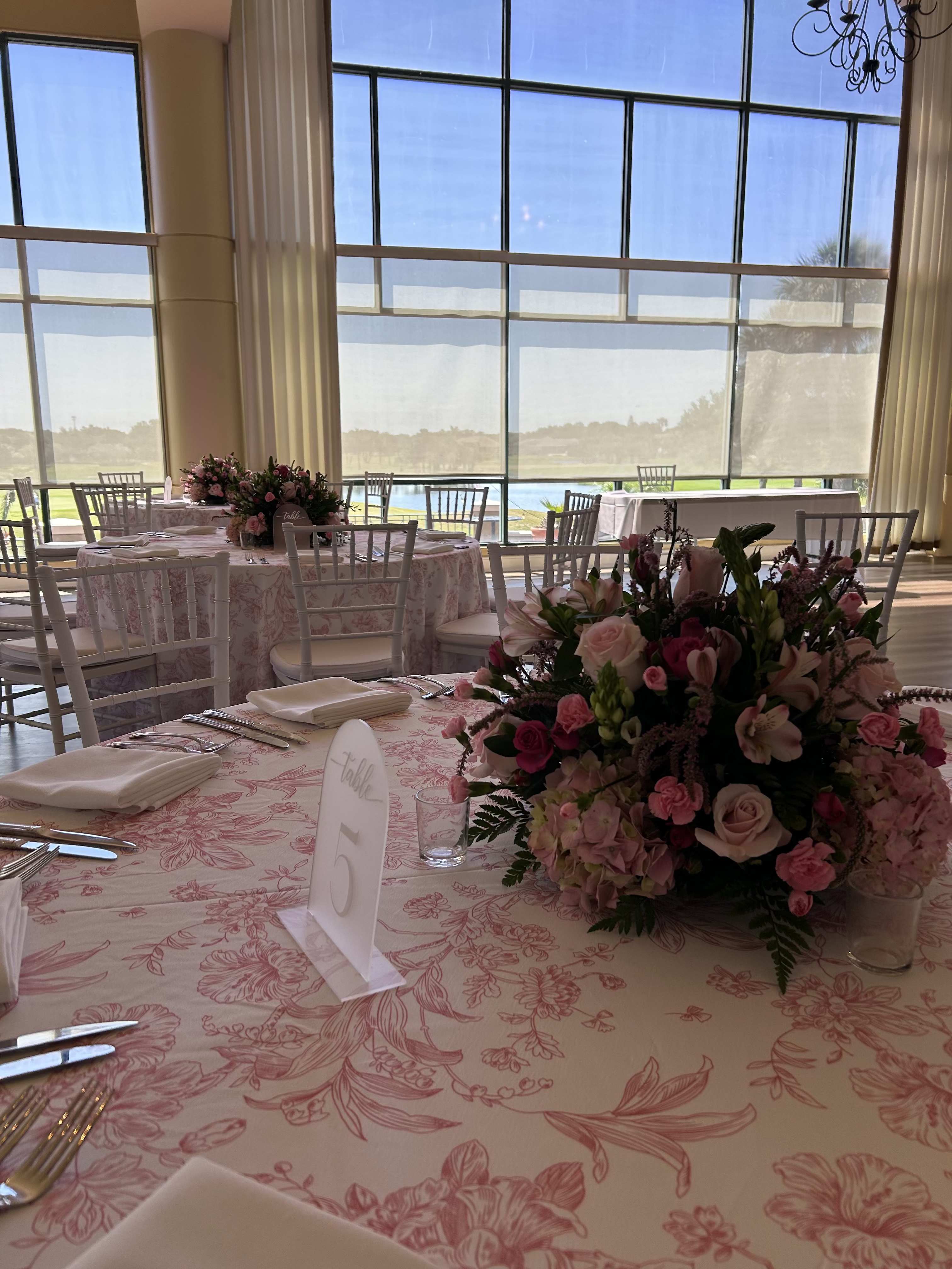 Lush low centerpiece  - A lush low centerpiece in soft pinks and whites, this wedding reception centerpiece mixes roses, lilies, alstroemeria, carnations and hydrangea-like clusters to create a layered, romantic focal point. Designed as a wedding or special event centerpiece, its compact, full shape keeps sightlines clear across the table so guests can talk easily while still enjoying the flowers' gentle scent and textural variety.   The arrangement pairs blush roses and pale lilies with the delicate speckled accents of alstroemeria, ruffled carnations, and rounded hydrangea clusters, punctuated by deeper green foliage for contrast and depth. The low profile and generous fullness make it ideal for round or long tables; in the photo it's styled with a pink floral tablecloth, folded white napkins, glassware and a table number card to reinforce an elegant reception look.  Perfect for brides seeking a soft, romantic palette, this centerpiece makes the person you're honoring feel celebrated and seen-gentle, classic, and decidedly festive. Because it's low and dense, it's easy to place at the center of guest tables; keep blooms fresh by changing the water every other day and removing any spent flowers, and order in sizes appropriate to your table dimensions to ensure the right proportion for your event.