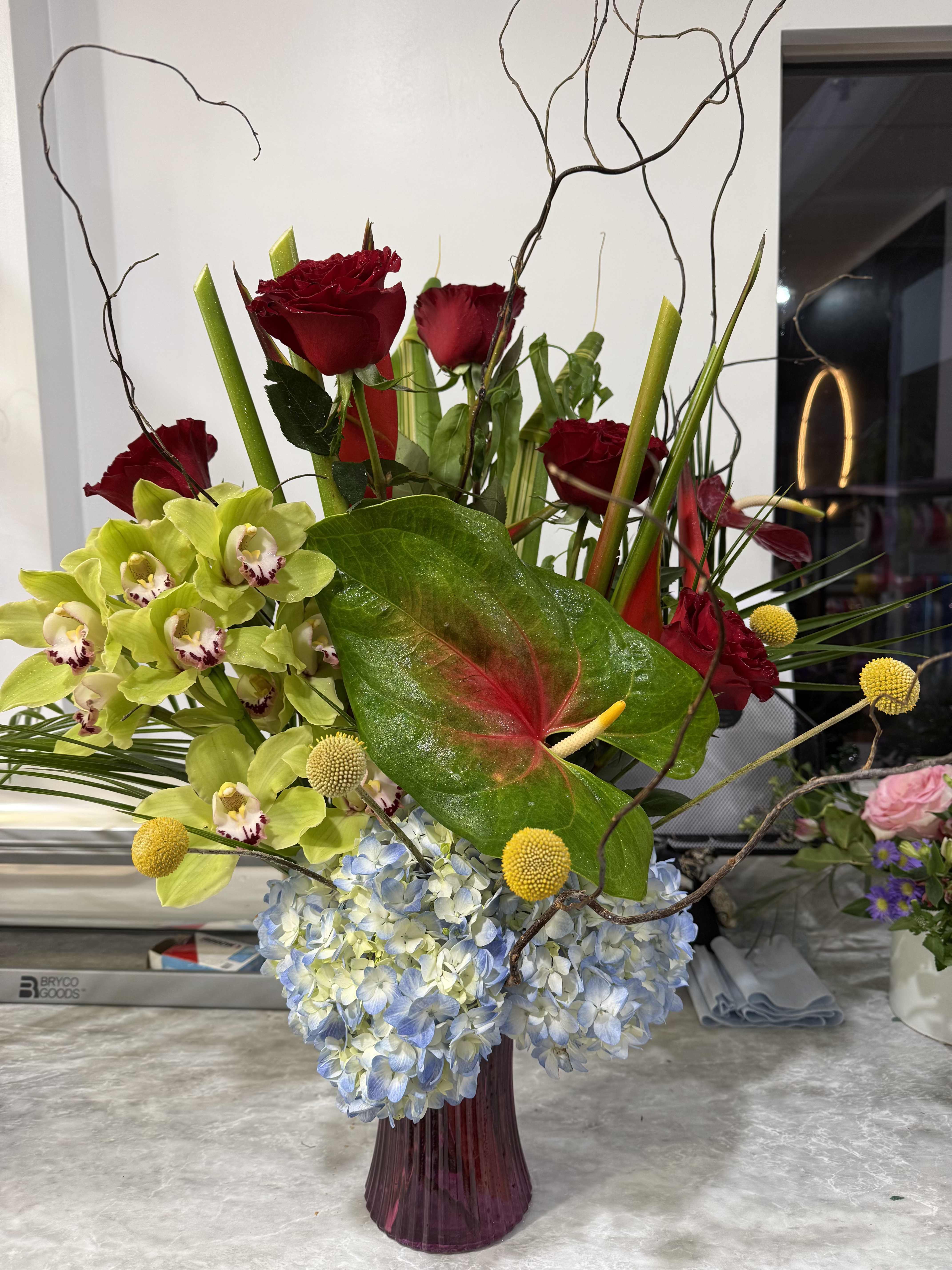 Exotic Tropical Beauty - Lush pale blue hydrangeas anchor this Exotic Tropical Beauty arrangement, surrounded by deep red roses and lime-green orchids for a bold, contemporary look. As a flower arrangement for a wedding reception, anniversary, or upscale celebration, it reads as dramatic and sculptural-the kind of centerpiece that makes the person you're honoring feel truly celebrated.  A tall purple glass vase holds a glossy anthurium leaf at the focal point, with thin curly branches rising above the composition to add height, movement, and architectural interest. Small yellow pom-pom accents punctuate the cooler blues and greens while the saturated reds bring warmth and contrast. The mix of roses, orchids, hydrangeas and complementary foliage creates varied textures-from the velvet petals of roses to the cloudlike hydrangea clusters and sleek orchid blooms.  Perfect for gifting to someone who loves modern, tropical style or for elevating an event table, this arrangement arrives ready to display in its vase; keep stems in fresh water and away from direct heat to extend bloom life. Choose this when you want a striking, made-for-the-moment floral statement that reads as both exotic and refined.
