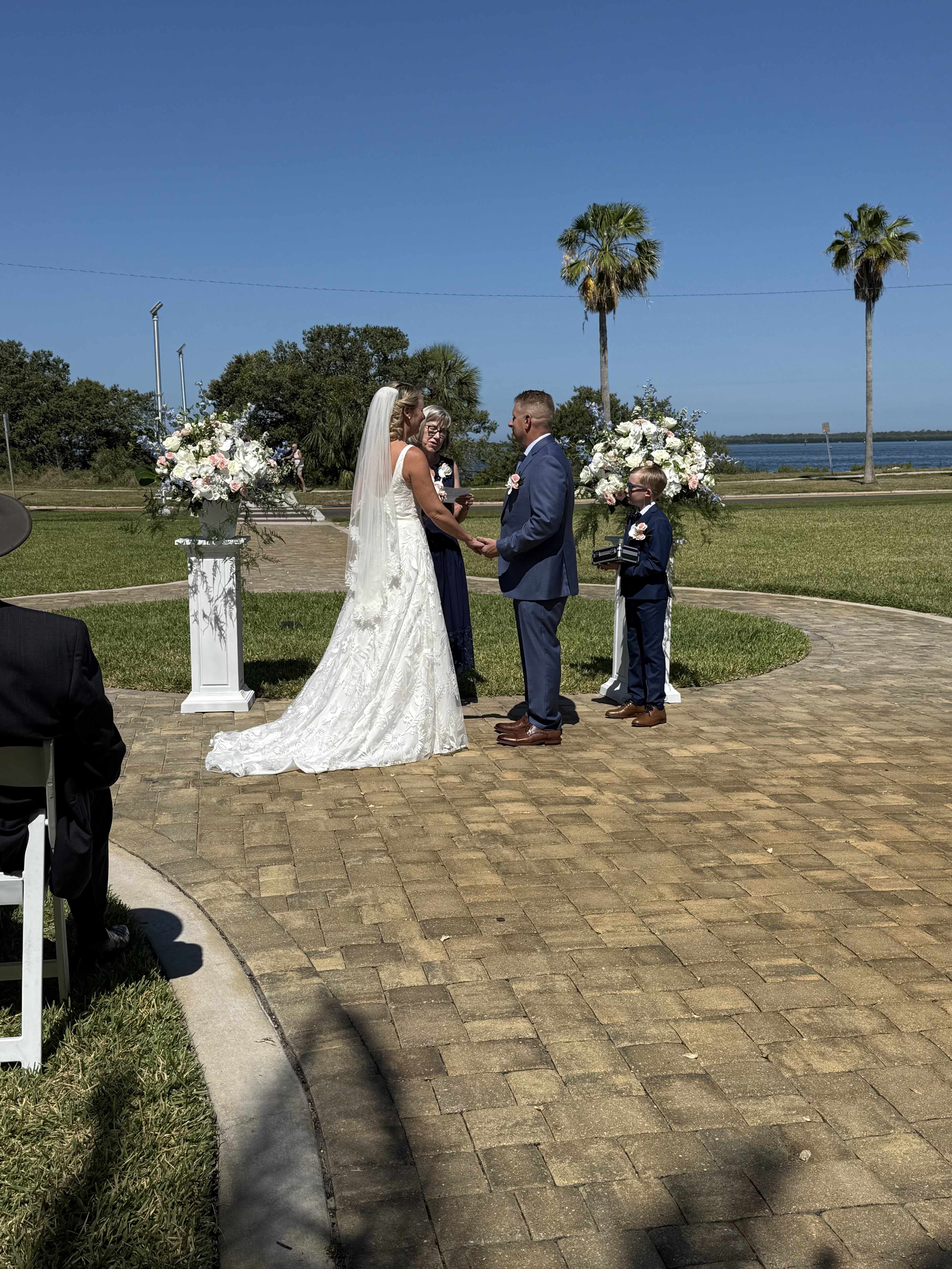 Elegant Ceremony Designs - Soft white roses and billowing hydrangeas rise from elegant white pedestal stands to frame the aisle-an ideal choice for flowers for weddings and outdoor ceremony décor. These Elegant Ceremony Designs give the couple a classic, romantic backdrop: predominantly white blooms warmed with soft pink accents and airy blue-purple filler that catch the breeze and read beautifully against a waterfront sky. The elevated compositions create a formal, celebratory presence that makes the ceremony feel deliberate and unforgettable.  Each arrangement pairs full-headed roses and hydrangeas with complementary greenery and delicate filler to add texture and depth; the color palette of white, pink, green, and blue keeps the look soft and airy. Shown on tall white pedestals beside bridal veil and wedding attire, these designs are scaled to command the altar without overpowering photographs. They're perfect for couples seeking timeless elegance-choose placement at the aisle entrance or flanking the couple to ensure your person feels truly celebrated on the day.  For an outdoor wedding, these pieces arrive ready to place on-site; discuss delivery and setup with our team to secure pedestal placement and final positioning. Light misting maintains freshness on warm days, and our florists tailor stem counts and scale to match small elopements or grand waterfront ceremonies so the arrangements fit your space and vision.