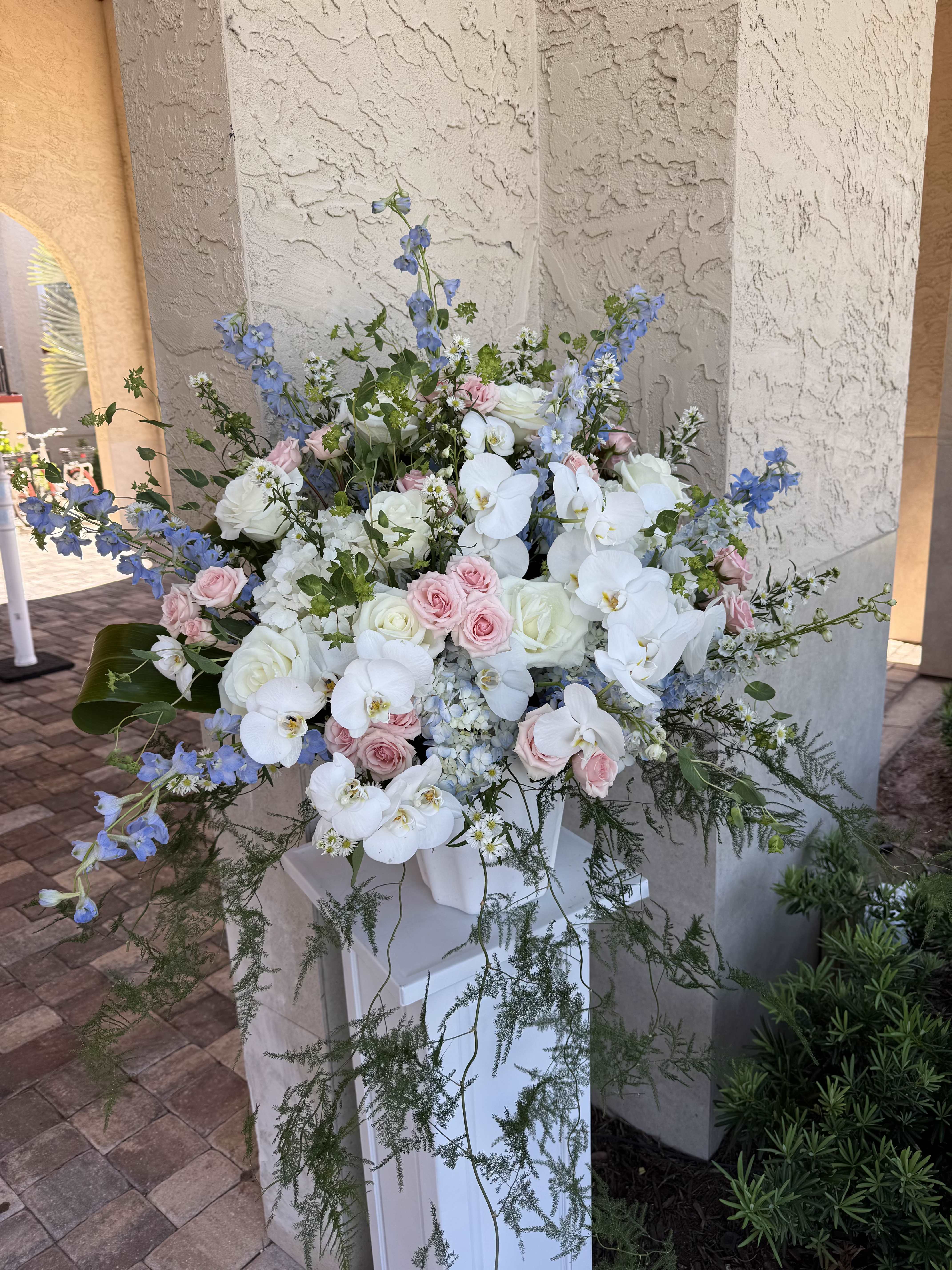 Phalaenopsis Orchid Ceremony Florals - White phalaenopsis orchids cascade gracefully from a tall, airy arrangement-perfect as ceremony florals or wedding centerpieces that need to read elegant from the aisle. This wedding flowers composition pairs those sculptural white orchids with pale pink roses and tall blue delphinium to create a romantic, garden-style statement that makes the person you're celebrating feel seen and cherished.  The design layers soft blue delphinium spikes above a mix of hydrangeas, daisies, and delicate white filler flowers for texture and depth, while trailing greenery brings movement and softness. Presented in a white pedestal vase, the arrangement stands tall and formal; the palette of white, pink, blue, and green reads fresh and refined for upscale celebrations.  Ideal for altar displays, entrance pieces, or paired along a long ceremony table, these florals elevate the moment without overpowering other décor. The pedestal vase makes placement simple-sizing and delivery details are available at checkout-care for the phalaenopsis and cut stems by keeping them cool and away from direct sunlight until the ceremony.