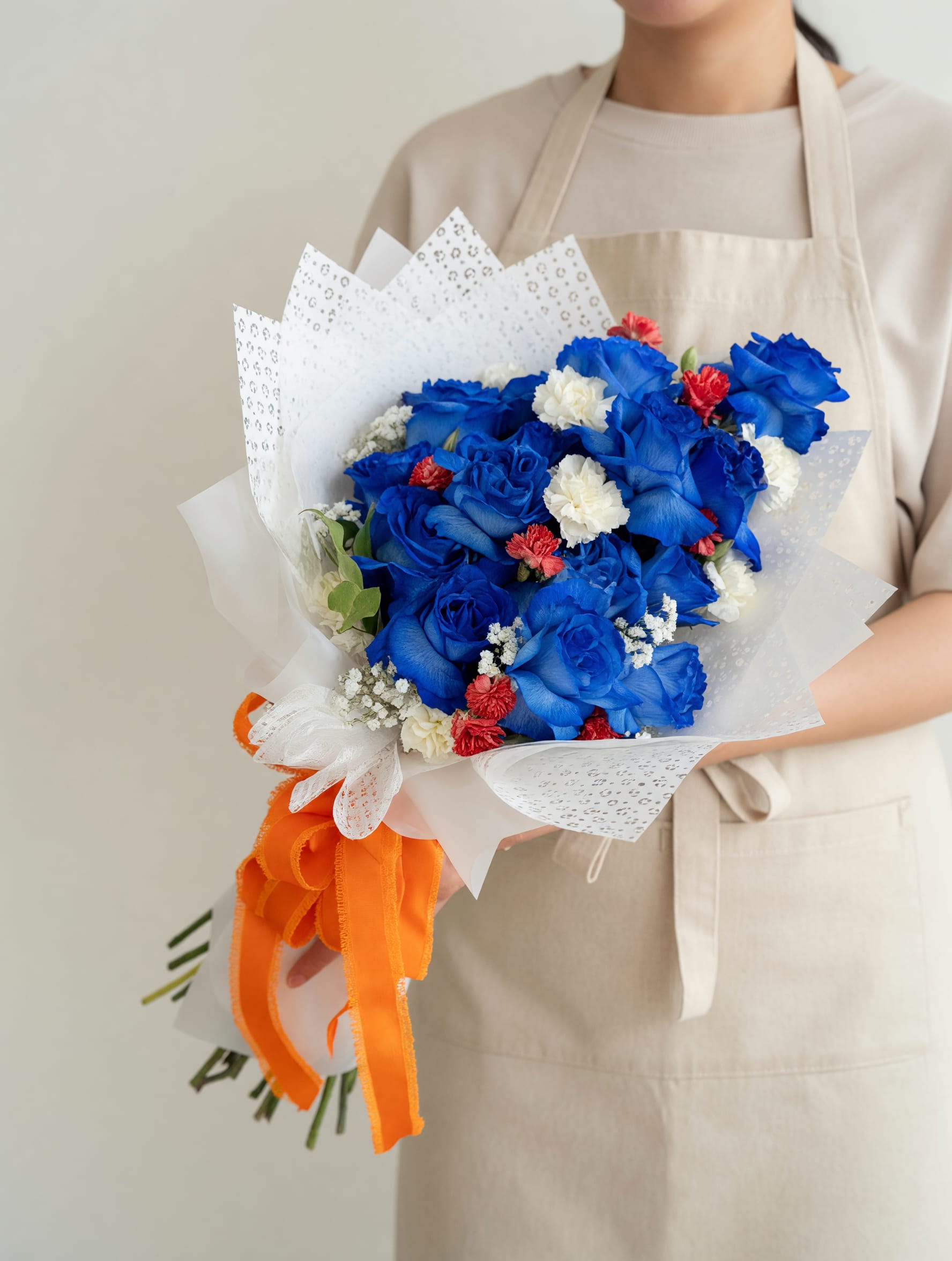 Birds Up! - Vivid blue roses take center stage in this celebratory bouquet-a bold flower arrangement perfect for graduation, school spirit gifts, or cheering on the Roadrunners. The blue roses are paired with small white filler blooms and pops of orange carnations to create a high-contrast, spirited look that reads festive and romantic at once-ideal for a UTSA grad, a proud alum, or anyone who roots for the University of Texas at San Antonio.  Tightly gathered and hand-tied, the bouquet is wrapped in layered white paper with a patterned outer wrap and finished with decorative white lace ribbon and a bright orange ribbon at the base for a modern, gift-ready presentation. Colors: blue, white, and orange. Flowers: roses, carnations, and complementary blooms for texture. This polished arrangement makes the recipient feel seen and celebrated-whether you're sending congratulations, school-day pride, or a thoughtful pick-me-up. Order with a short note to make it unmistakably for UTSA spirit.