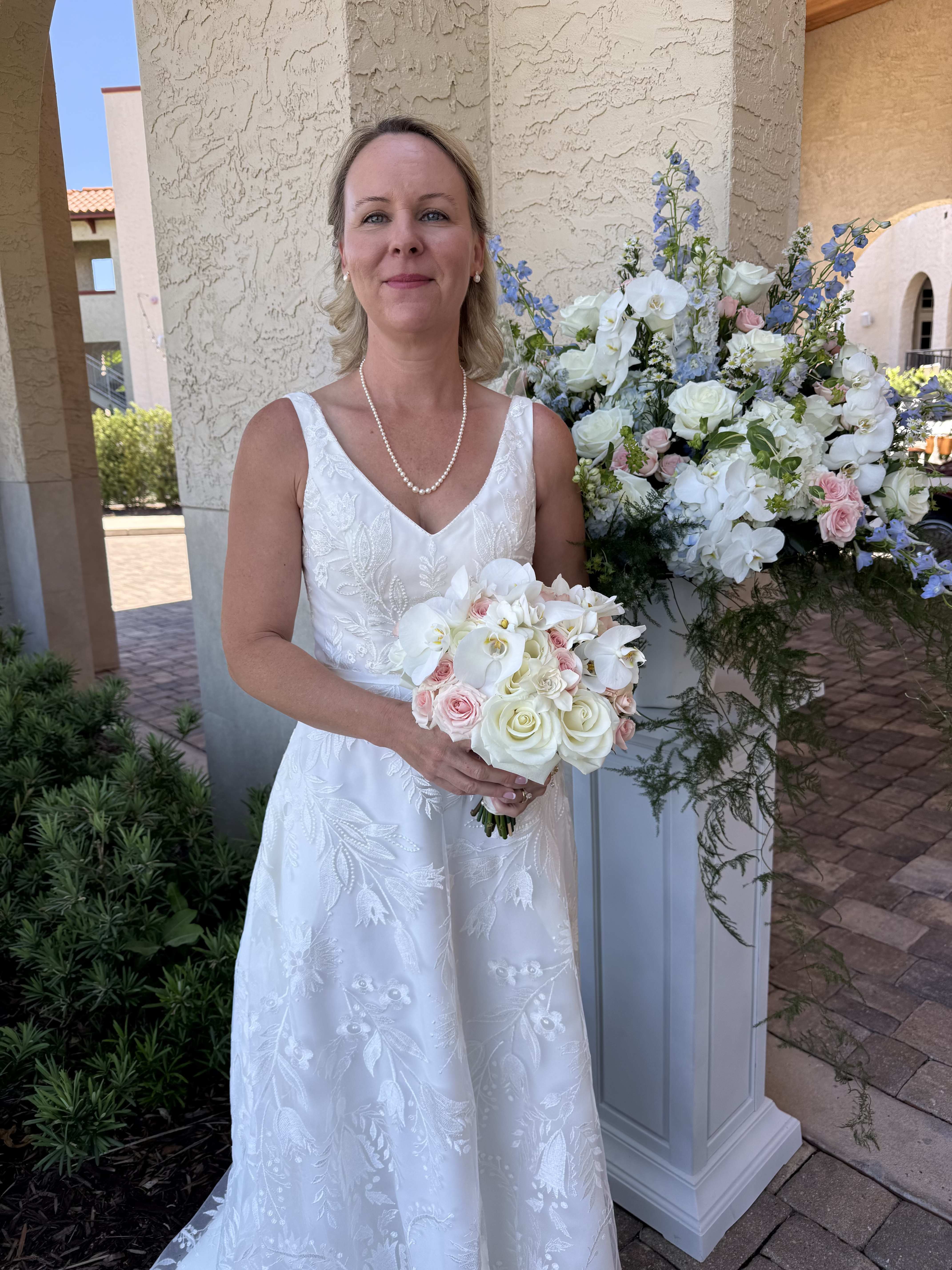 Blushing Romance Bridal Bouquet - A soft pastel palette of white roses, gardenia, blush spray roses, and white Phalaenopsis orchids creates the kind of romantic wedding bouquet that makes the bride feel gracefully radiant-perfect as a bridal bouquet for a ceremony or as a gift for the newlyweds. The rounded, medium-sized bouquet pairs with a larger coordinating floral pedestal stand to bring continuity from aisle to reception; tall blue delphinium accents add vertical drama and a whisper of cool contrast against the soft pinks and greens.  This arrangement features classic roses and delicate orchids joined by airy delphinium and cheerful daisies, with fresh greenery for texture and movement. The bouquet's compact, hand-held shape sits beautifully with bridal details like a wedding dress and pearl necklace, while the larger pedestal arrangement uses the same flowers to create a formal focal point that complements ceremony backdrops or sweetheart tables.  Choose this for a bride who prefers timeless elegance with a modern pastel twist-ideal for spring or summer ceremonies and intimate receptions. The medium bouquet is easy to carry down the aisle, and the coordinating pedestal piece elevates your floral décor without overwhelming the space; order soon to ensure flower availability and on-time delivery for your wedding date.