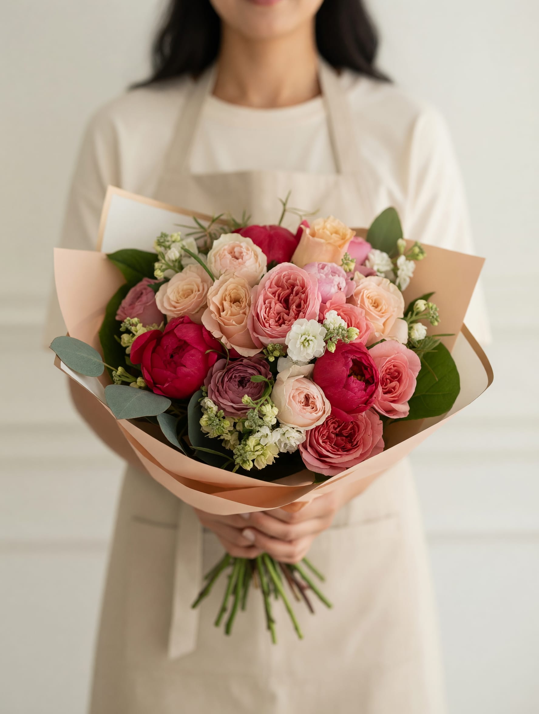 Coral Girl - Layered roses in blush, peach, coral and deep pink form the heart of this hand-tied flower arrangement, making Coral Girl an instantly romantic birthday bouquet or anniversary surprise. The tightly clustered roses and spray roses read lush and full, while small white filler blooms and rounded, eucalyptus-like greenery give a soft, fresh contrast that keeps the look light and celebratory.  Wrapped in pale blush and brown paper with stems hand-tied for a natural, gift-ready finish, this bouquet mixes roses, spray roses, carnations and textured blooms for a rich, dimensional feel. The palette of pink, peach, white and green suits Birthdays, I Love You, or "Just Because" moments - the kind of gift that makes the person you're thinking of feel truly seen. Care is simple: keep stems in fresh water and trim them slightly on arrival to extend vase life; sizes are hand-tied, so expect a full, garden-inspired presentation.