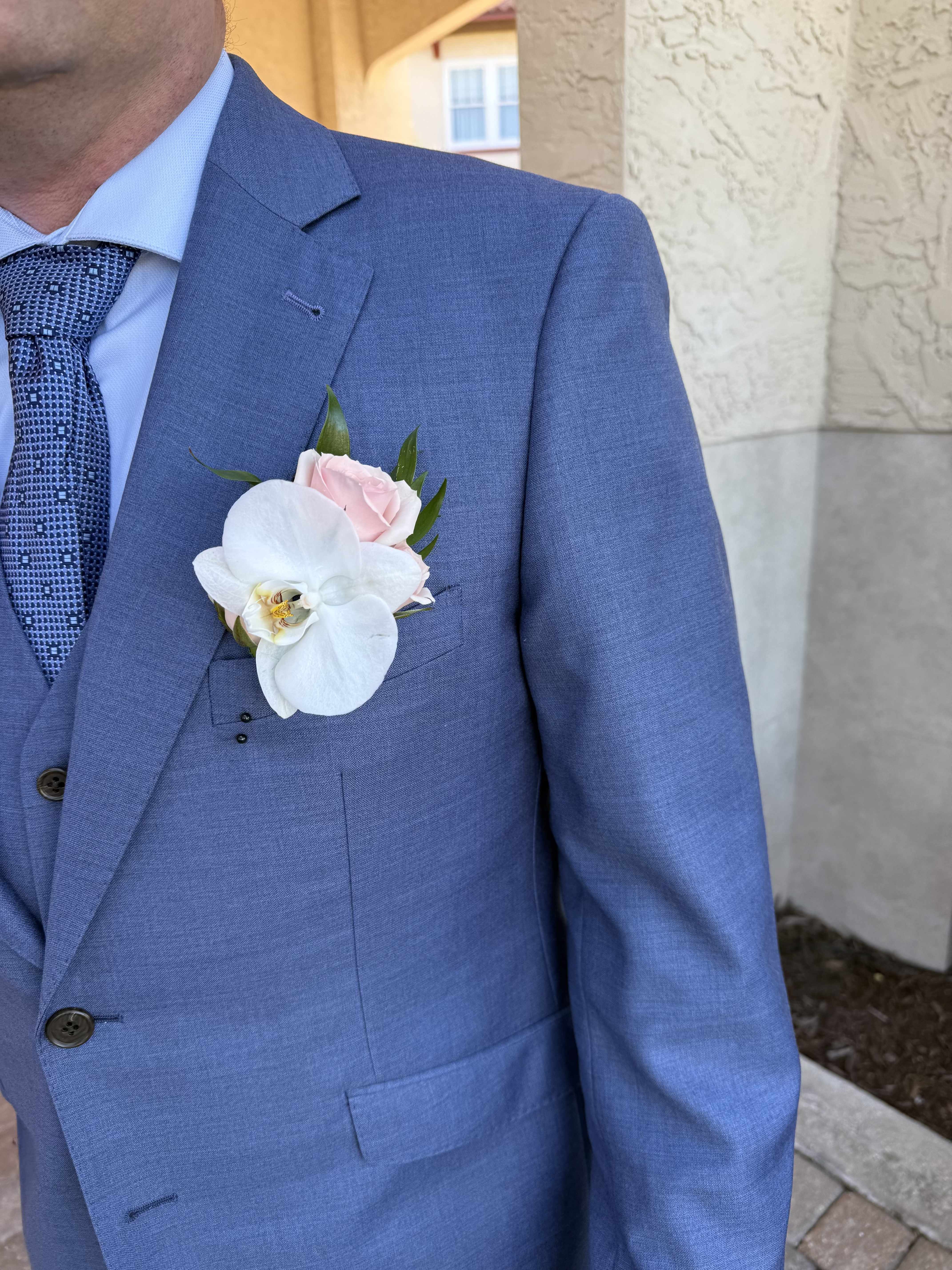 Classic Orchid Boutonniere - A single white Phalaenopsis orchid bloom anchored by a soft pink rose creates an elegant focal point for weddings and formal occasions-perfect as a groom, groomsman, or father-of-the-bride boutonniere. The white-and-blush palette reads romantic and refined, while the compact, polished design sits neatly on a blue suit jacket to complete a tailored ceremony look.  This Classic Orchid Boutonniere pairs the smooth, sculptural texture of the orchid with the velvety warmth of a pink rose and small green accents for freshness and contrast. Its restrained size keeps the arrangement formal and wearable without overwhelming lapels; the secure pinning and low-profile build ensure it stays in place throughout vows, photos, and dancing.  Made to coordinate with bridal bouquets and wedding party attire, this boutonniere is a meaningful finishing touch that will make the person you're thinking of feel celebrated and seen. For best results, keep blooms cool and dry before placing on the suit; select same-day delivery or pickup to preserve peak freshness.