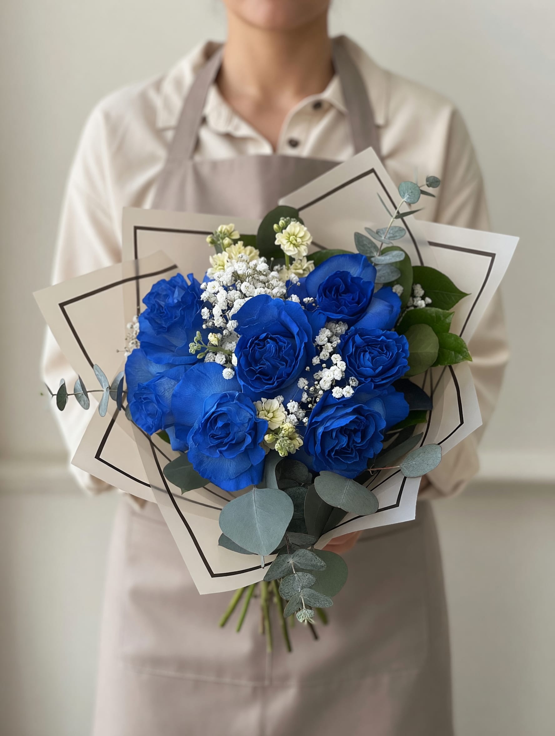 Something Blue - Vivid blue roses take center stage in this hand-tied flower arrangement, making an immediate, modern statement for a birthday bouquet, anniversary roses, or a romantic congratulations gift. The cool blue palette paired with soft white and pale cream accents creates a look that feels both romantic and slightly unconventional - the kind of bouquet that will make the person you're thinking of feel truly special and seen.  Tightly clustered blue roses are complemented by small white filler blooms and rounded eucalyptus sprigs that add airy texture and a fresh green contrast. Wrapped in layered cream or translucent gray paper with dark trim and finished with a glossy satin ribbon, the presentation is crisp and polished; decorative wrapping paper and thoughtful apron-style paper wrap give the bouquet a gift-ready finish. The cool-toned combination reads dramatic yet elegant, perfect for someone who loves contemporary floral design.  Ideal for celebrating birthdays, engagements, congratulations, or simply saying "I'm thinking of you," this arrangement arrives ready to present and makes a memorable impression. Treat the recipient to a bouquet that balances bold color with soft accents - no special care beyond fresh water is needed to enjoy its layered textures and long-lasting visual impact.