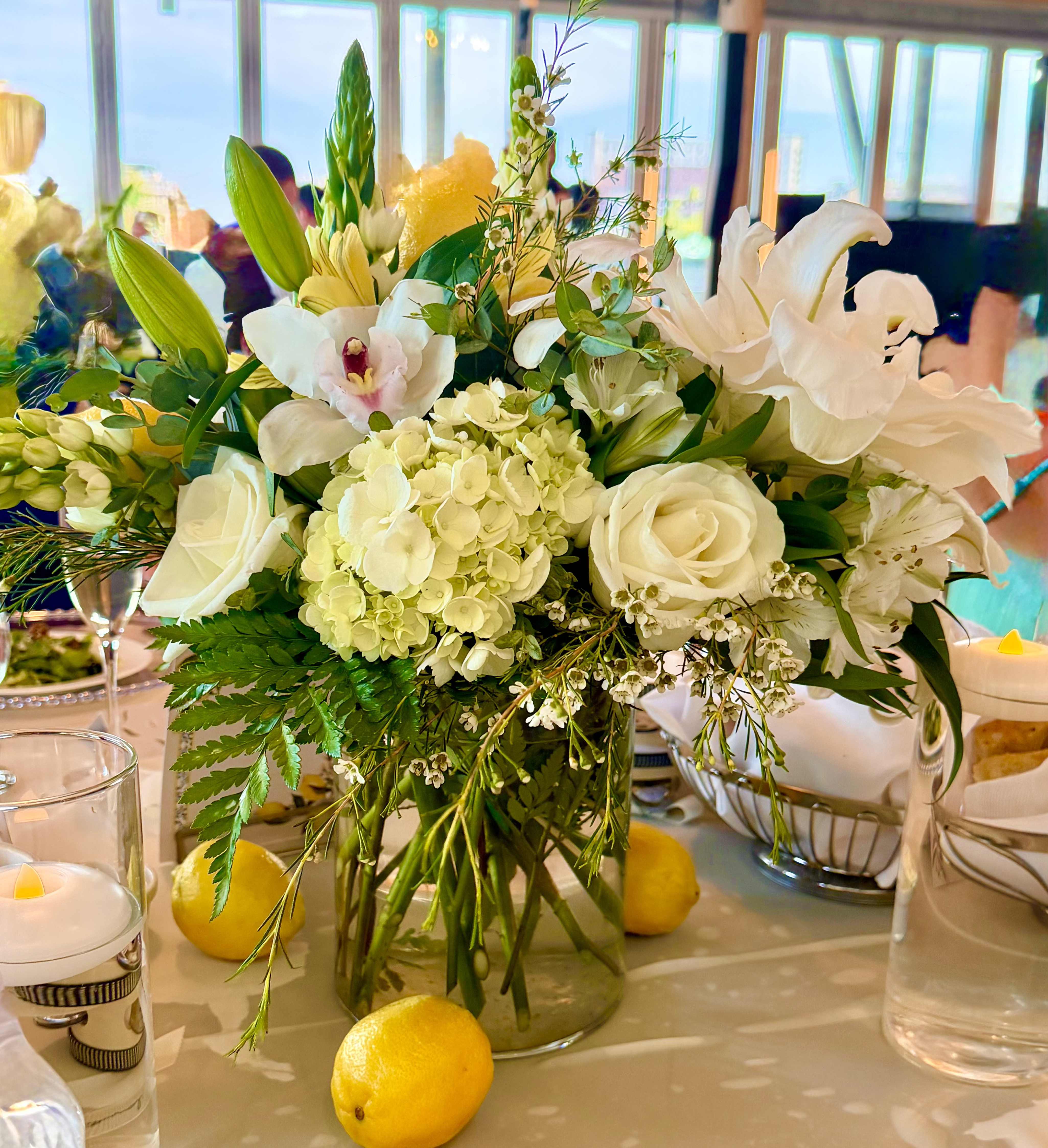 Lemon Garden Centerpiece - Bright clusters of white hydrangeas and roses anchored in a clear glass vase give this wedding centerpiece its lush, garden-fresh presence-perfect for spring ceremonies or receptions. The Lemon Garden Centerpiece pairs white lilies and delicate orchid accents with trailing greenery and airy branches, creating a soft, romantic silhouette that reads elegant from every table. Placed among glass candle holders and pale candles, the arrangement becomes a glowing focal point that makes the person you're celebrating feel cherished and celebrated.  This centerpiece combines large, layered hydrangea blooms with white roses and lilies, finished with orchid touches and varied green foliage for texture and movement. Fresh lemons tucked into the design add a bright yellow pop against the clean white palette, reinforcing a cheerful, refined aesthetic. Arranged in a clear glass vase and shown with glass candle holders and lit candles, it suits long banquet tables or intimate round settings-bringing a fragrant, natural look with a polished event-ready finish.