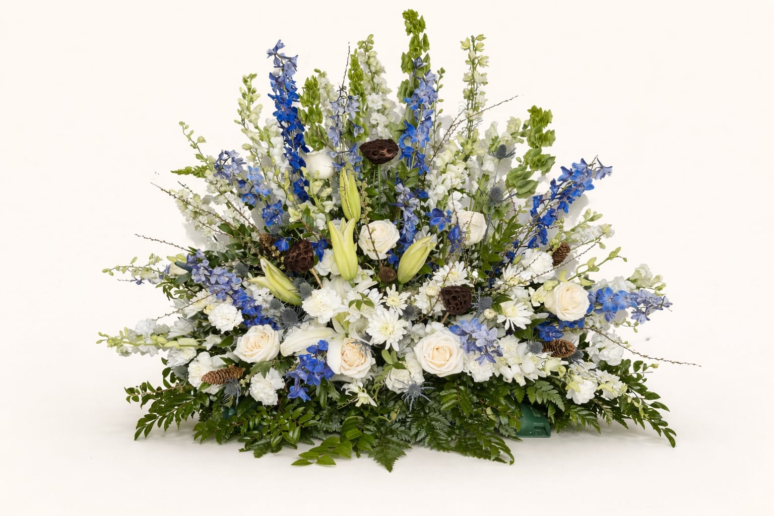 Serenity in Blue - A cool, dignified sympathy spray built to offer comfort: this large, low fan-shaped funeral arrangement blends white roses and lilies with vertical blue delphinium and clusters of white daisy-like blooms to create a serene blue-and-white palette ideal for memorial services. The design's dense center and taller vertical accents give a graceful, reverent silhouette that reads beautifully at the front of a casket or on an easel for a service.  Crafted on a floral foam base, the piece features layered greens and a few pinecones for natural texture and subtle rustic contrast. Expect a substantial, floor-level presentation with a full, compact center and taller stems rising behind-flowers include roses, lilies, delphinium, daisies, and chrysanthemums in white, blue, and green tones.  Choose this arrangement when you want the person you're remembering to feel honored and seen; it conveys sympathy and calm without being showy. Because it's built in floral foam for event display, order with service timing in mind so the flowers arrive at peak freshness for the ceremony.