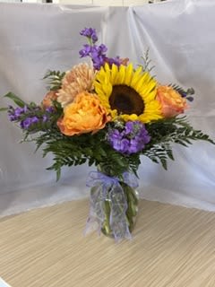 Grey House Blooms-All That Jazz - A bold yellow sunflower takes center stage in this cheerful flower arrangement-perfect as a birthday bouquet, a get-well surprise, or an everyday gift to lift someone's spirits. The mix of sunflowers, chrysanthemums, and smaller accent blooms in bright yellow, tangerine orange, purple, and fresh green reads joyful and energetic; it's the kind of arrangement that makes the person you're thinking of feel instantly noticed and smiled at.  The design has a rounded, hand-tied silhouette that balances full, textured blossoms with delicate filler for a lively, garden-fresh look. It arrives in a clear glass vase tied with a sheer purple ribbon for a casual but polished presentation that suits both home and office. Ideal for sending happy vibes, this arrangement is crafted to look abundant from every angle and is sized to be a prominent tabletop focal without overwhelming a room.