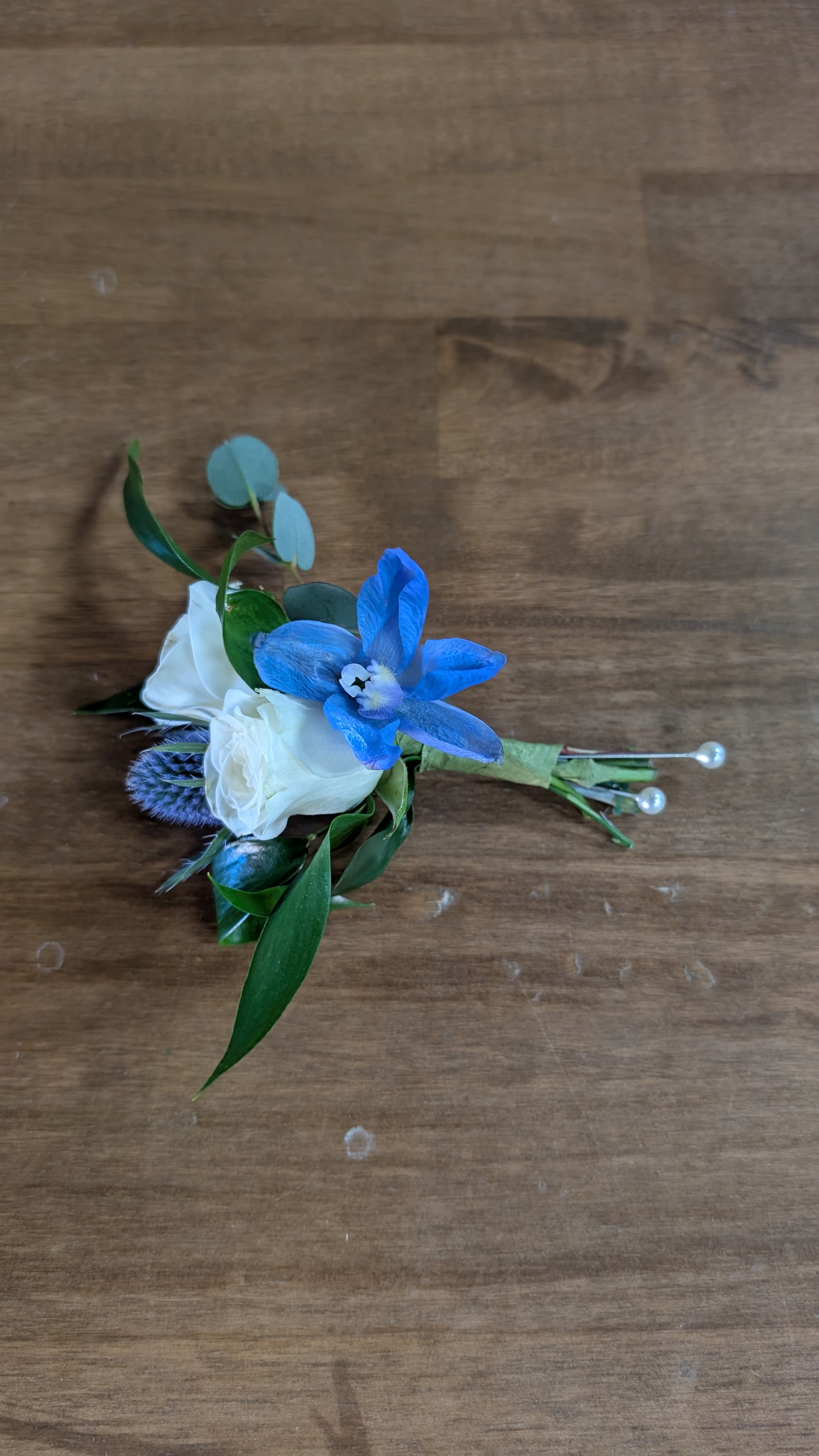 Boutonniere - A crisp blue orchid anchors this boutonniere, set beside two delicate white rose blooms for a fresh, modern take on wedding and formal boutonniere styling. Perfect for a groom, groomsman, or anyone attending a dressy event, the bright blue-and-white contrast reads elegant and intentional-the kind of detail that makes the person you're honoring feel thoughtfully dressed and noticed.  Glossy green leaves and a few round eucalyptus-style sprigs frame the flowers, keeping the silhouette compact and wearable. The stems are neatly wrapped with floral tape and secured with decorative pearl-tipped pins for a polished finish that holds through the day. Order as shown for a classic boutonniere that pairs especially well with navy, charcoal, or white suits; keep it cool and dry until wear and pin it on just before stepping out for best presentation.