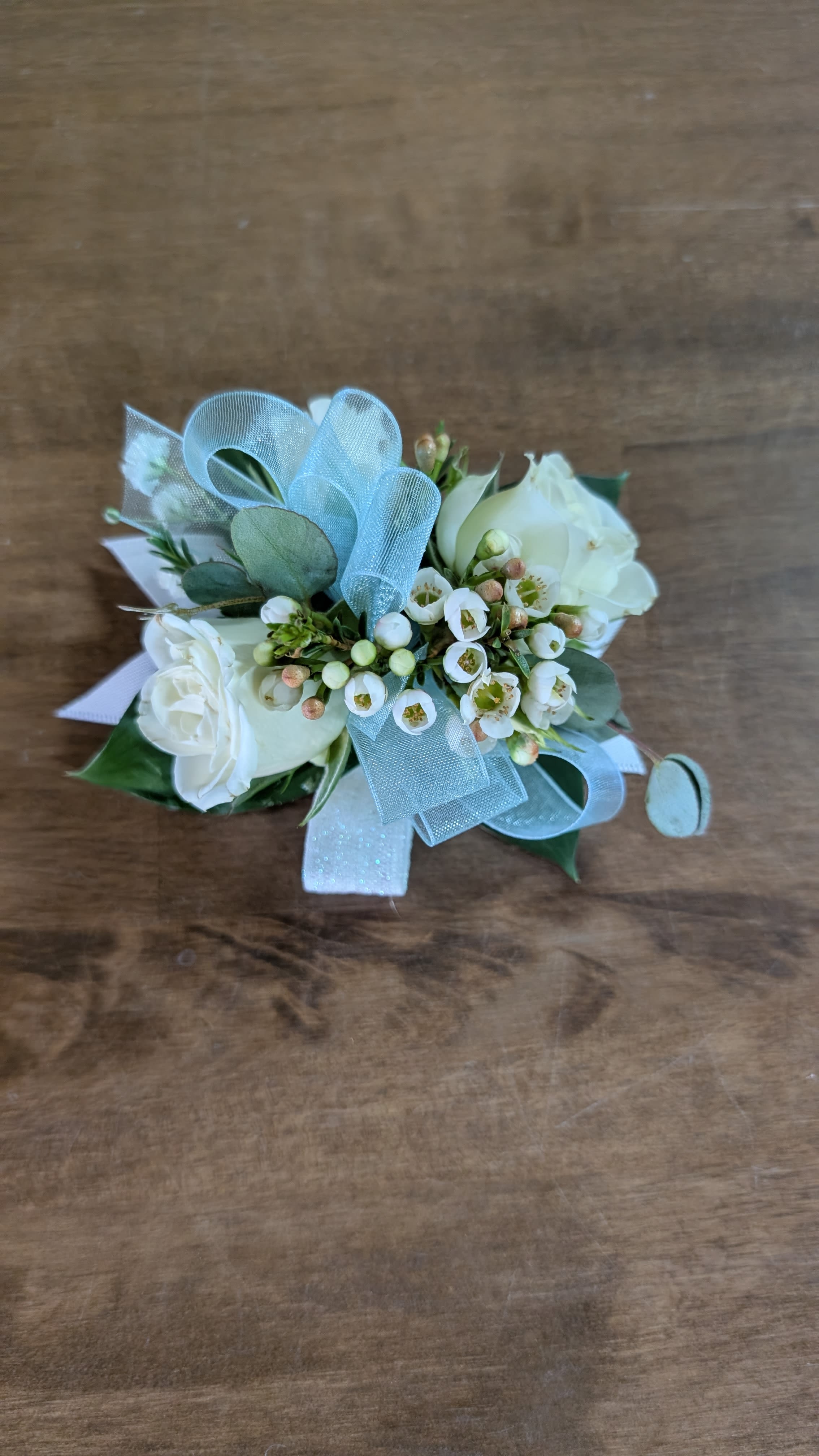 Corsage - A petite wristlet-style corsage arranged with roses and clusters of tiny filler blooms, tied with sheer ribbon loops for a soft, formal accent-perfect as a wedding corsage or prom accessory that feels fresh and airy. The compact, handcrafted shape reads as neat and feminine, designed to make the person wearing it feel truly celebrated on their special day.  Handcrafted with fresh roses, delicate filler flowers, decorative wrap and coordinating ribbons for a refined presentation, this small floral piece is sized to be comfortably worn on the wrist or pinned to a dress. Ideal for bridesmaids, prom dates, mothers of the bride, or anyone you want to honor; order with your event timing in mind to ensure peak freshness, store cool and mist lightly until wear.