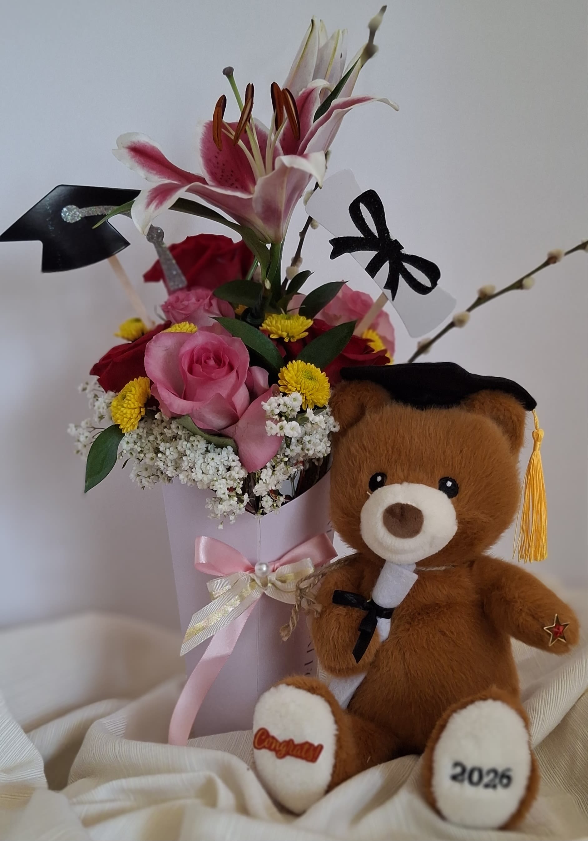 Scholar Bear &amp; Blooms - The Summa Cum Laude Bloom is a soft and sophisticated celebration of achievement. This arrangement features a stunning pink lily rising above a graceful blend of blush roses and cheerful yellow accents.