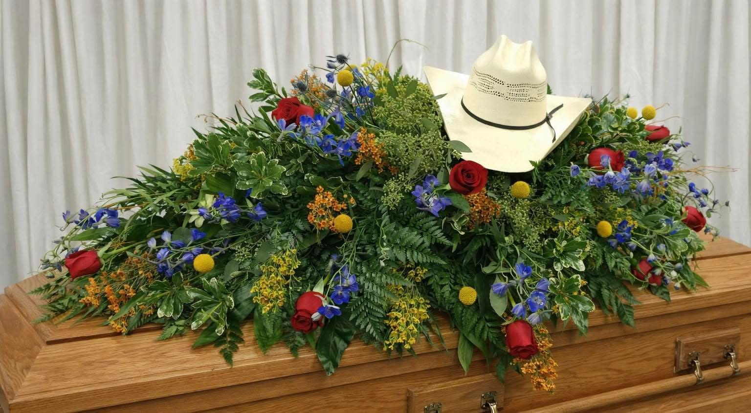 Gary Fields of glory - A bold, personalized casket spray that reads like a proud farewell: this large funeral flower arrangement combines red roses with vivid blue blossoms, yellow accents, and dense greenery to create a full, layered tribute for a memorial service. As a casket spray for a funeral or celebration of life, it communicates respect and individuality-especially fitting when the person you're honoring loved Western style or had a ranching background.  Laid low and wide across a wooden casket, the spray mixes classic red roses with other bright blue and yellow florals set into rich green foliage for depth and texture. A white cowboy hat is placed near the center as a heartfelt focal point, lending a distinctly Western character that makes the arrangement feel like a true reflection of the deceased's personality.  Choose this casket spray when you want a colorful, dignified memorial that feels personal-a meaningful way to say goodbye and celebrate a life lived with spirit. Because it's made to lie across a full-size casket, sizing and placement are arranged by our funeral delivery team to ensure a secure, elegant presentation; please contact us with service details so we can coordinate delivery and set-up.   If any design elements are of major importance to your order, please include them in the florist instructions at checkout or contact us to ensure availability.