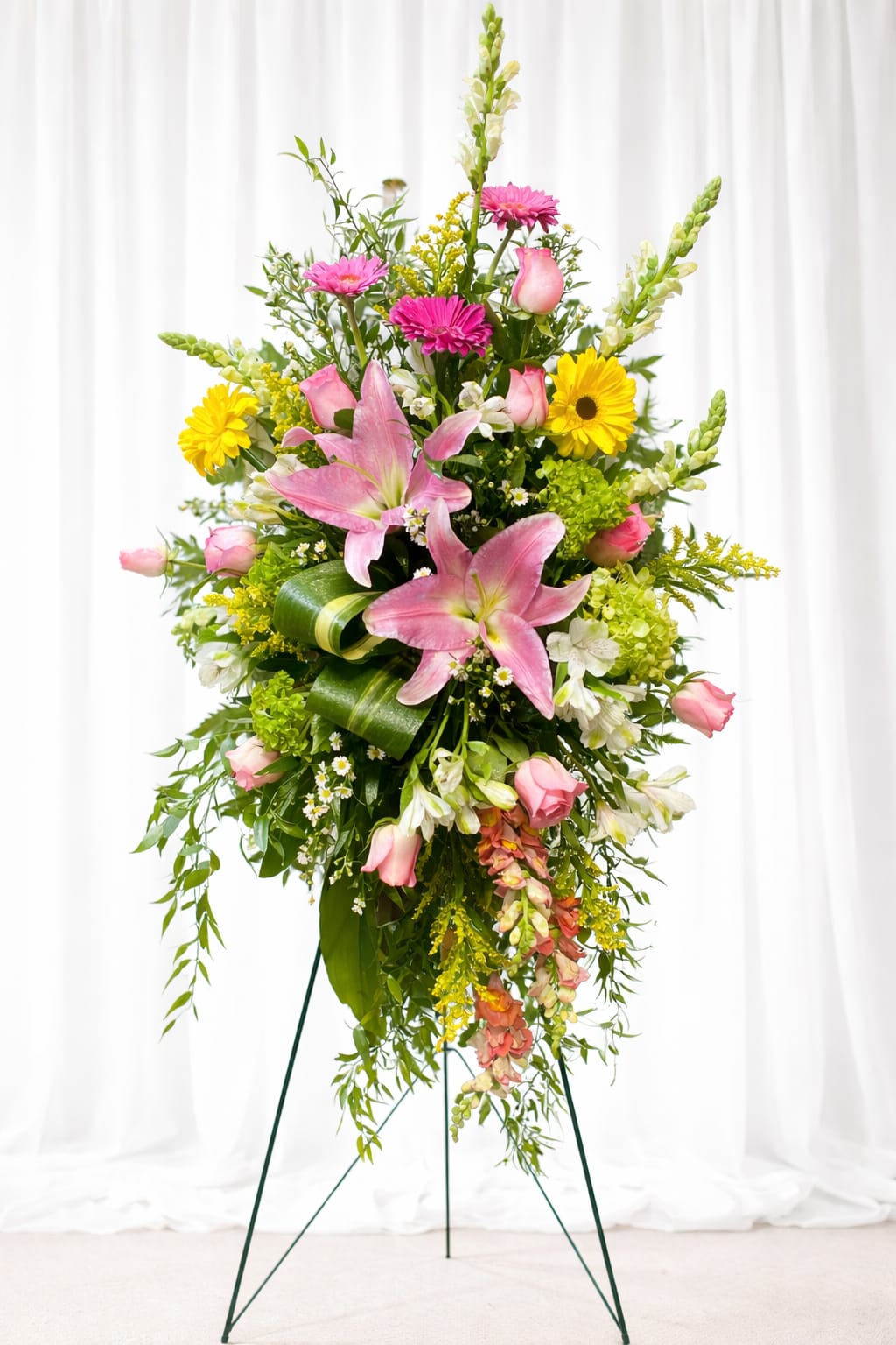 Heavenly blossoms - Tall pink lilies and roses rise above bright yellow daisy-like blooms in a graceful standing spray designed for sympathy and memorial services. As a funeral flower arrangement on an easel, this floral tribute reads with respectful presence-its soft pinks, whites, and touches of green convey warmth and remembrance while the upright composition and vertical snapdragon spikes lend a formal, ceremonial silhouette.  The arrangement features large pink lilies and pink roses paired with gerbera-style yellow flowers, white accents, chrysanthemums, snapdragons, and lush cascading greenery for texture and movement. Mounted on a floral easel stand, the piece is full and asymmetrical with a layered, vertical profile that fills sightlines at a service or reception. Sized to serve as a focal spray, it arrives ready to display on the included stand.  Made to comfort the person you're honoring, this arrangement offers a bright yet respectful expression that helps family and friends feel seen and remembered. Care is simple-place on the easel in a cool, sheltered spot away from direct heat; our florists prepare and secure the design so it's delivery-ready for the ceremony or memorial viewing.   If any design elements are of major importance to your order, please include them in the florist instructions at checkout or contact us to ensure availability.