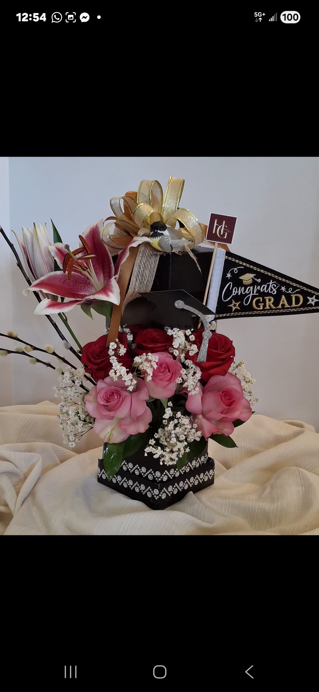Honor &amp; Triumph Bouquet - A sophisticated graduation masterpiece presented in an embellished black gala box. This premium arrangement blends red and pink roses with striking stargazing lilies and delicate baby's breath. Beautifully detailed with a festive "Congrats Grad" pennant flag, a glittery mini mortarboard cap, and a majestic gold bow featuring a keepsake songbird accent.