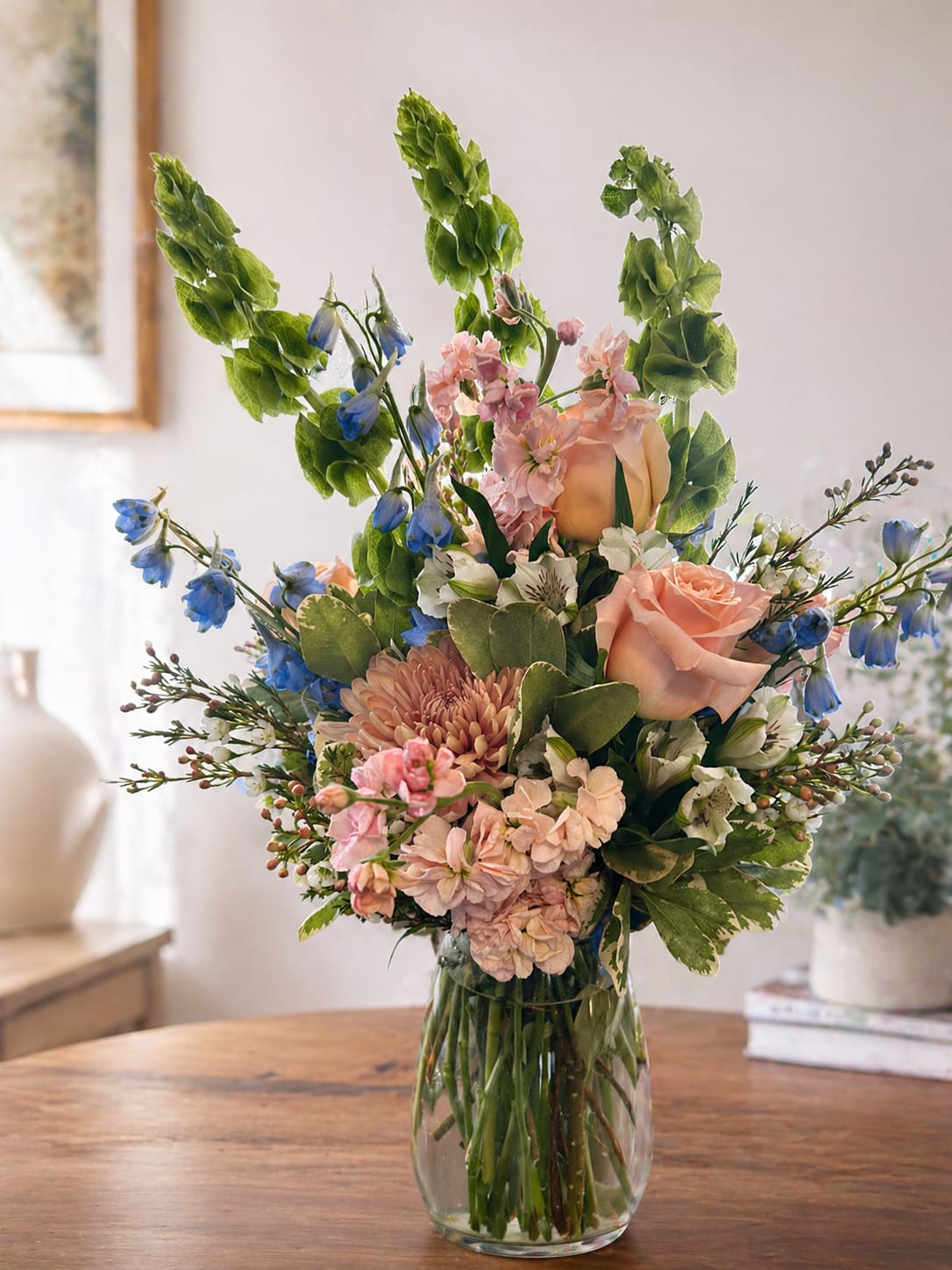 Peachy - Soft peach roses and pink daisy-like blooms mingle with delicate blue bell-shaped flowers in a fresh garden-style arrangement-perfect as a spring birthday bouquet or a thoughtful general gift arrangement. The airy, upright silhouette creates a sense of gentle movement that reads romantic without feeling fussy, making it an ideal choice when you want the person you're thinking of to feel light, cherished, and noticed.  This flower arrangement features peach roses, pink daisy-like flowers, alstroemeria and white accent blooms arranged with tall green stems and upright branching elements for a loose, vertical shape. All stems are presented in a clear glass vase that highlights the natural texture and colors-soft peach, blush pink, blue, and verdant green-for an elegant, seasonal look.  Made to celebrate birthdays, springtime moments, or simply to send a warm thinking-of-you message, this bouquet brings a fresh, garden feel to any room. Arrange on a table or bedside dresser; change the water every 2-3 days and trim stems slightly to extend vase life for several days of enjoyment.   If any design elements are of major importance to your order, please include them in the florist instructions at checkout or contact us to ensure availability.