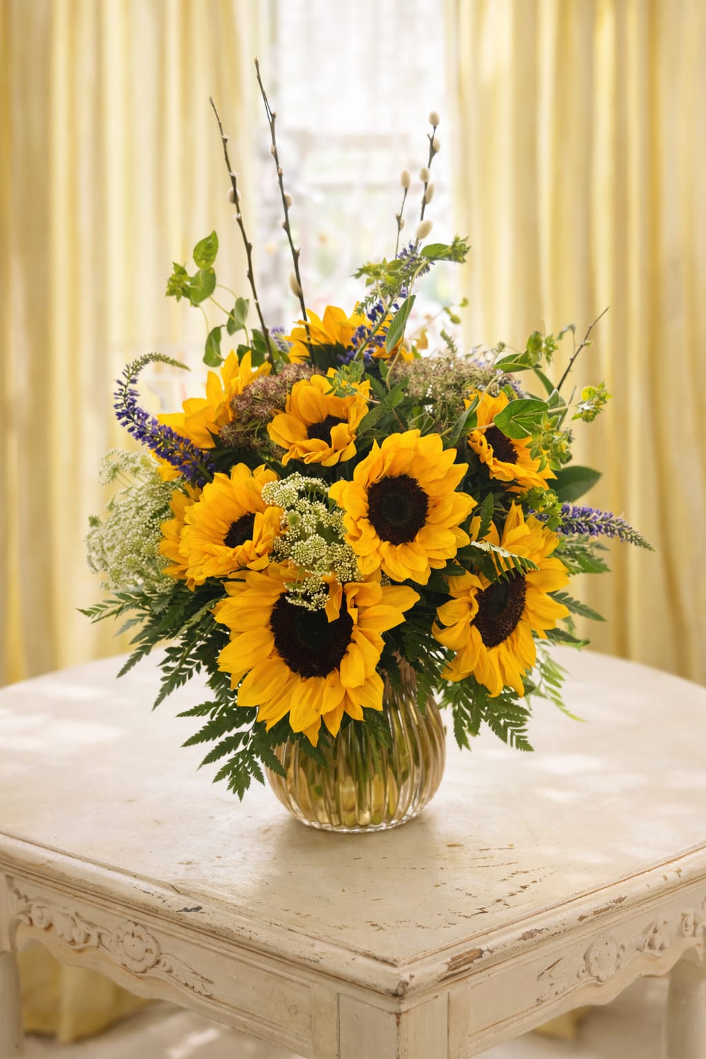 Sunrise  - Bright, golden sunflowers take center stage in this cheerful flower arrangement-a sunny bouquet perfect for birthdays, get-well wishes, or just because you want to brighten someone's day. The arrangement reads like a fresh-picked garden with tall purple accent spikes and delicate white filler blooms surrounding the bold yellow heads, so the person you send it to will feel instantly uplifted and noticed.  Set in a simple clear ribbed glass vase, the loose, garden-style composition uses airy greenery to give the bouquet a full, natural silhouette that works well on a kitchen table or office desk. The mix of yellow, green, purple, and white creates warm seasonal energy without feeling fussy, making it a versatile choice for everyday gifting or summer celebrations.   If any design elements are of major importance to your order, please include them in the florist instructions at checkout or contact us to ensure availability.