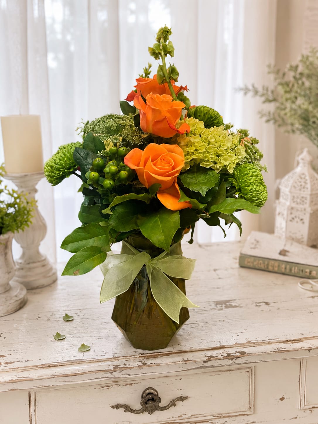Sunshine elegance - Vivid orange roses take center stage in this cheerful flower arrangement, giving the recipient an instant lift-perfect as a birthday bouquet, congratulatory gift, or bright everyday surprise. The lively mix reads as a celebratory arrangement and will make the person you're thinking of feel noticed and genuinely cheered.  Clustered with green and yellow textural blooms and soft white accents, the garden-style design is full and upright with a tall central stem and rounded side blooms for layered depth. Arranged in a faceted glass vase and finished with a pale ribbon bow, it arrives ready to display on a table or desk and brings a sunny, modern touch to any room.  Ideal for sending joy to a friend, coworker, or family member celebrating a milestone, this piece balances bold color with fresh greenery so it fits spring and summer occasions-or anytime you want to brighten someone's day. Standard vase size holds the arrangement securely; refresh water every couple of days and trim stems slightly to extend vase life.   If any design elements are of major importance to your order, please include them in the florist instructions at checkout or contact us to ensure availability.