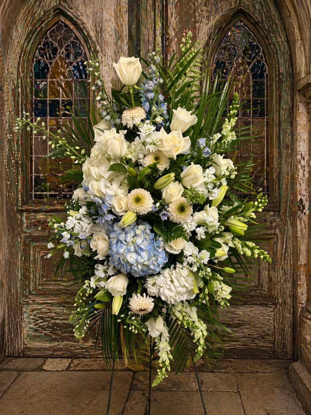 Winter blues  - A cool, dignified standing spray designed to comfort and honor-the Winter blues sympathy arrangement brings serene white and pale blue tones to memorial services and graveside farewells. This tall standing floral tribute uses formal, architectural lines to convey respect and calm, making it an appropriate choice to express sympathy or to mark a memorial occasion.  Dense layers of white roses, lilies, hydrangea, chrysanthemums and snapdragons are accented with soft blue hydrangea and pale blue delphinium-like spikes for a restrained, wintery palette. Long palm fronds and other linear greenery frame the oval composition on a standing easel, giving the piece a structured, elegant silhouette that reads well from a distance and up close.  Made to be displayed on a standing easel, this spray is sized for funeral services and memorial displays; its classic shape and soothing colors help the person you're honoring feel remembered and seen. For arrangements intended for a specific service time, please include delivery instructions at checkout so we can ensure timely placement.   If any design elements are of major importance to your order, please include them in the florist instructions at checkout or contact us to ensure availability.