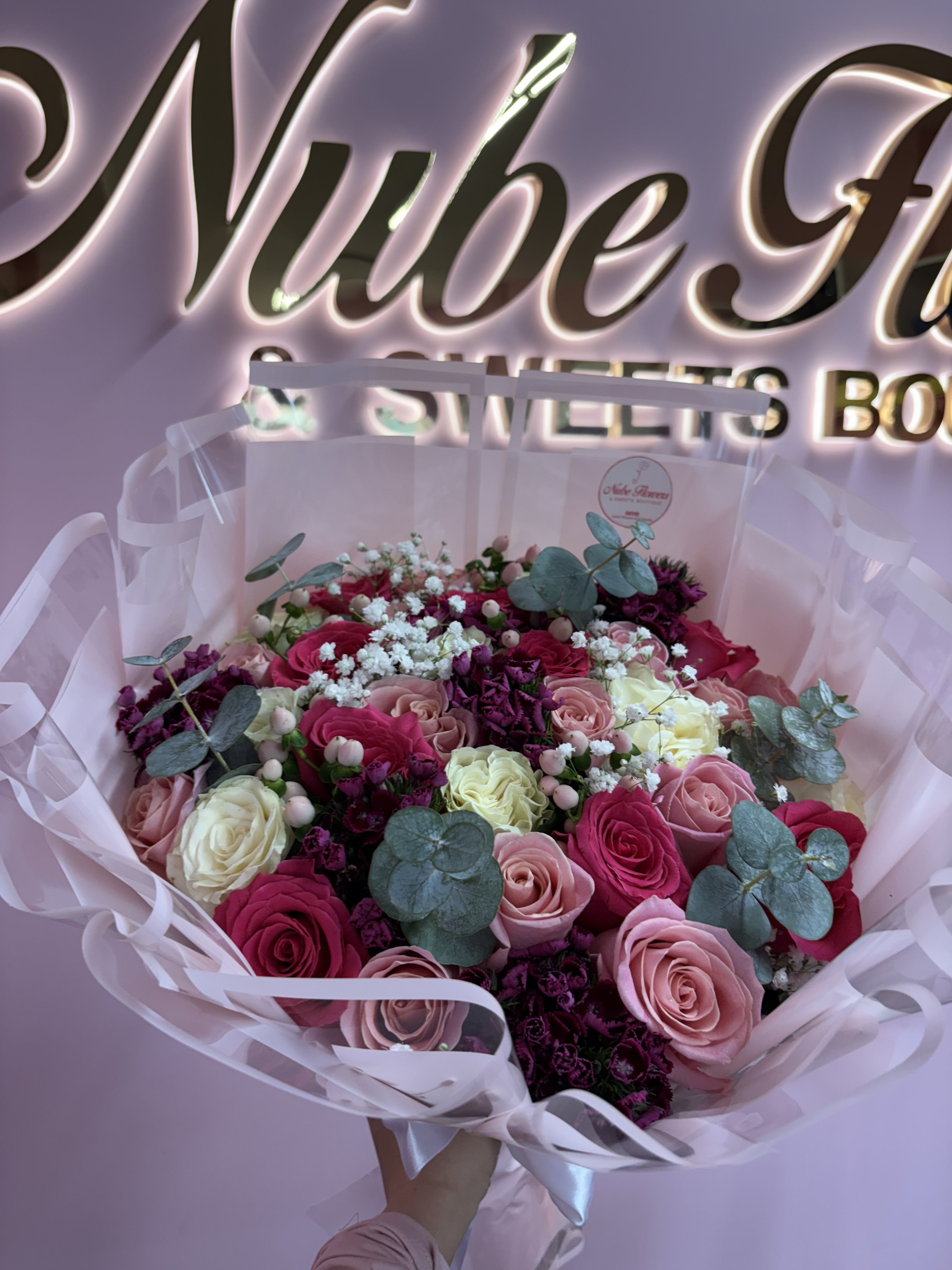 Mix Blooms - A lush hand-tied flower arrangement of tightly packed roses in hot pink, blush, cream and white makes a joyful centerpiece for anniversaries, birthdays, or Valentine's Day. The Mix Blooms bouquet reads romantic and celebratory-designed to make the person you're thinking of feel truly seen, adored, and celebrated the moment it arrives.  This bouquet combines roses with small white filler blooms and deep purple accent flowers woven through cool blue-green, eucalyptus-like foliage for contrast and texture. It's wrapped in soft pastel decorative paper and finished with a satin ribbon for an elegant, gift-ready presentation. The round, abundant shape feels full and luxurious rather than airy or minimal.  Ideal for someone who loves classic romance with a modern color twist, this arrangement arrives ready to display-no fussing required. To keep blooms looking their best, trim stems and refresh the water every couple of days; ordering options include standard hand-tied sizing to suit the occasion.