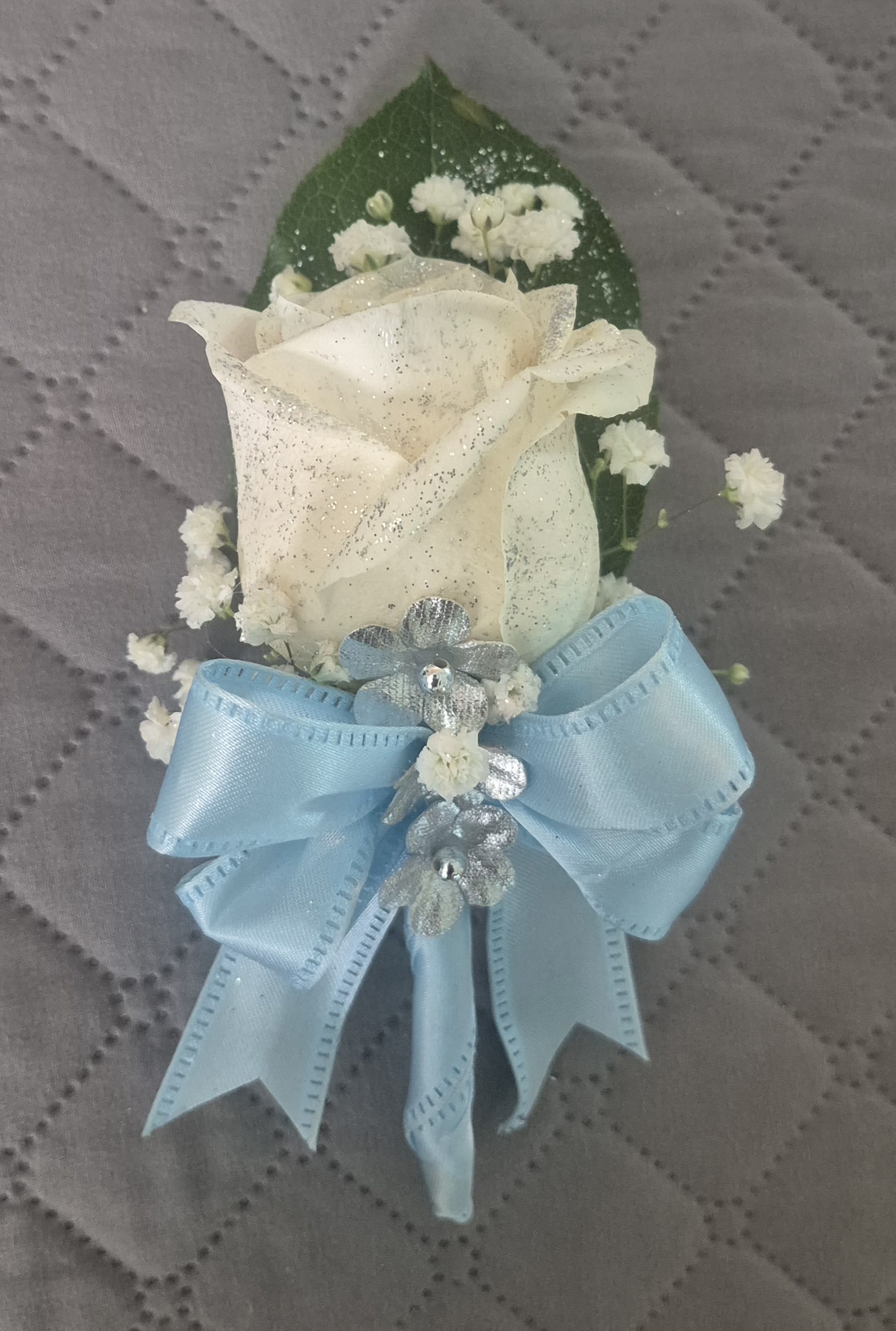 WINTER WHISPE ROSE BOUTONNIERE - AN ELEGANT WHITE ROSE BOUTONNIERE ACCENTED WITH DELICATE BABY'S BREATH, SHIMMERING SILVER DETAILS, AND A SOFT BLUE SATIN BOW. A TIMELESS FLORAL PIECE PERFECT FOR WEDDINGS, PROMS, BAPTISMS, OR ANY SPECIAL CELEBRATIONS.