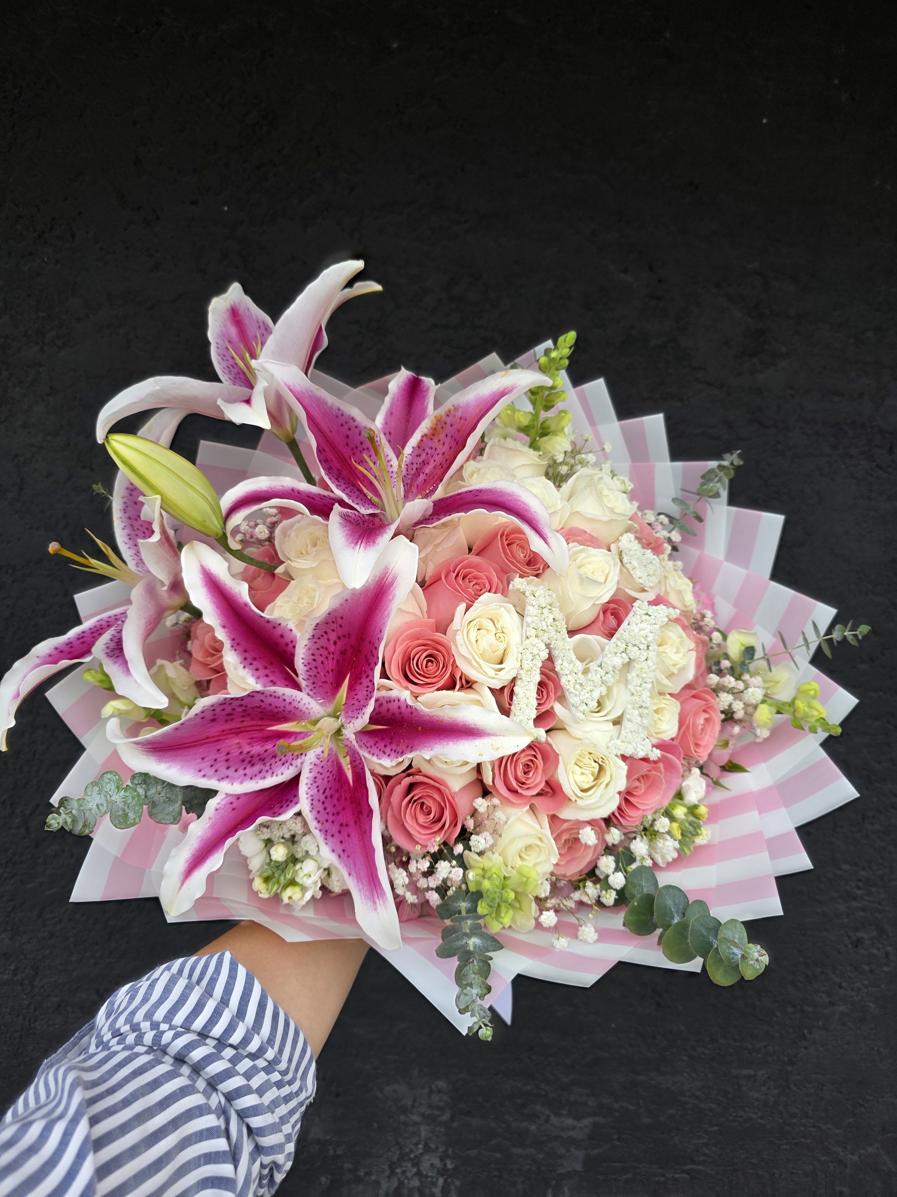 Love You Lots Bouquet- 50 Roses  - Velvety pink and ivory roses mingle with large pink-and-white oriental lilies in a lush, hand-tied flower arrangement that reads as romantic celebration - perfect as an anniversary bouquet, Mother's Day gift, birthday surprise, or a "just because" way to say I love you. The layered pink-and-white striped wrapping and neat white ribbon give this floral gift a polished, present-ready look that will make the person you're thinking of feel truly adored and seen.  This arrangement features fifty roses in pink and ivory tones, accented by showy lilies, delicate white filler blooms, and soft eucalyptus-like foliage for gentle texture and scent. The design is full and round with a generous profile; it can also be delivered in the included glass vase for an elegant display. Colors stay firmly in the romantic pink, white, and green palette for a feminine, celebratory impression.  Choose this bouquet when you want the recipient to feel cherished - whether you're marking a milestone, saying thank you, or brightening someone's day. For best results, trim stems and change water every two days; lilies may open further over the first 24-48 hours to increase fragrance and drama.