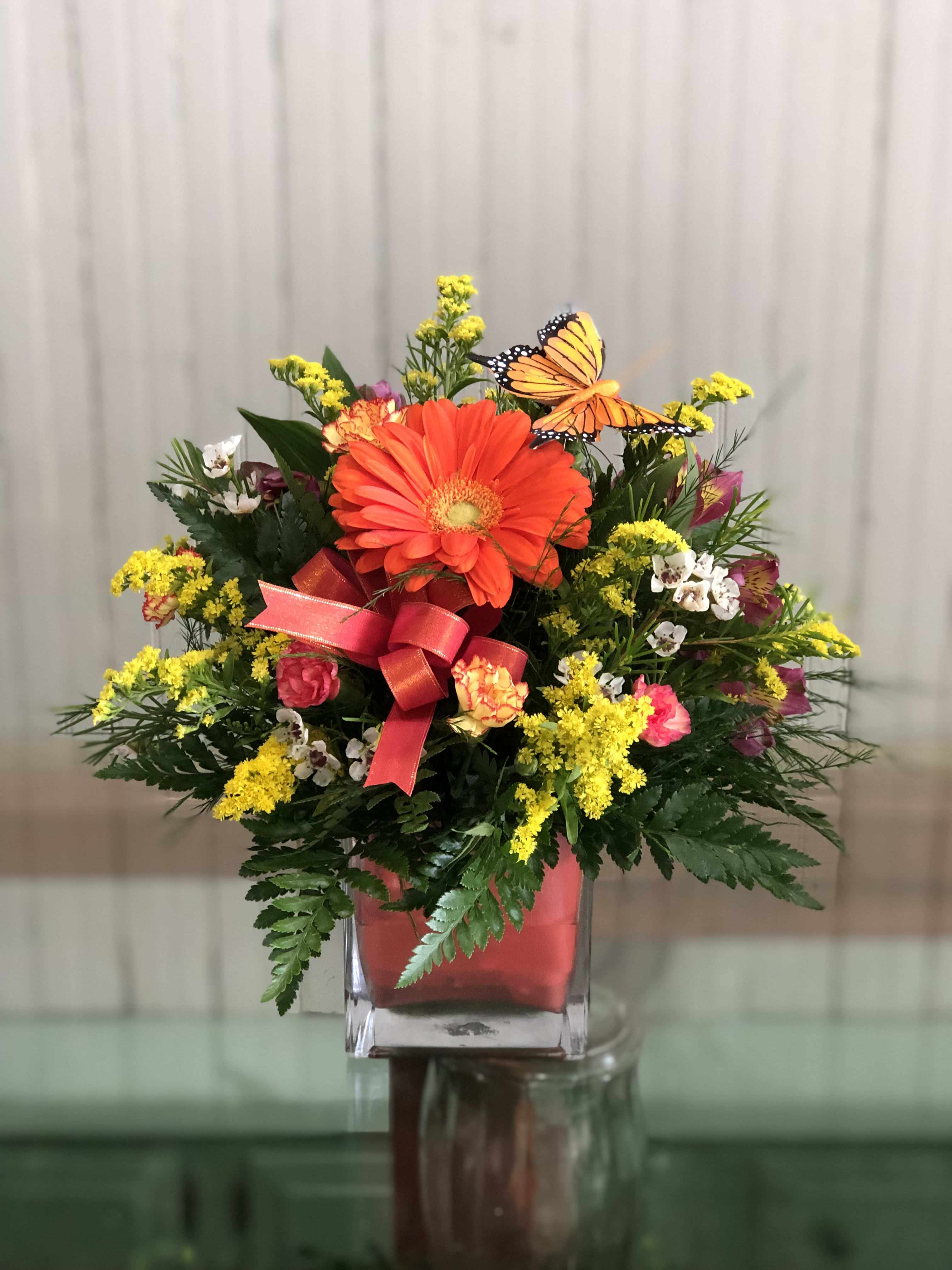 Bright mixed flower arrangement in a square glass vase with a butterfly accent