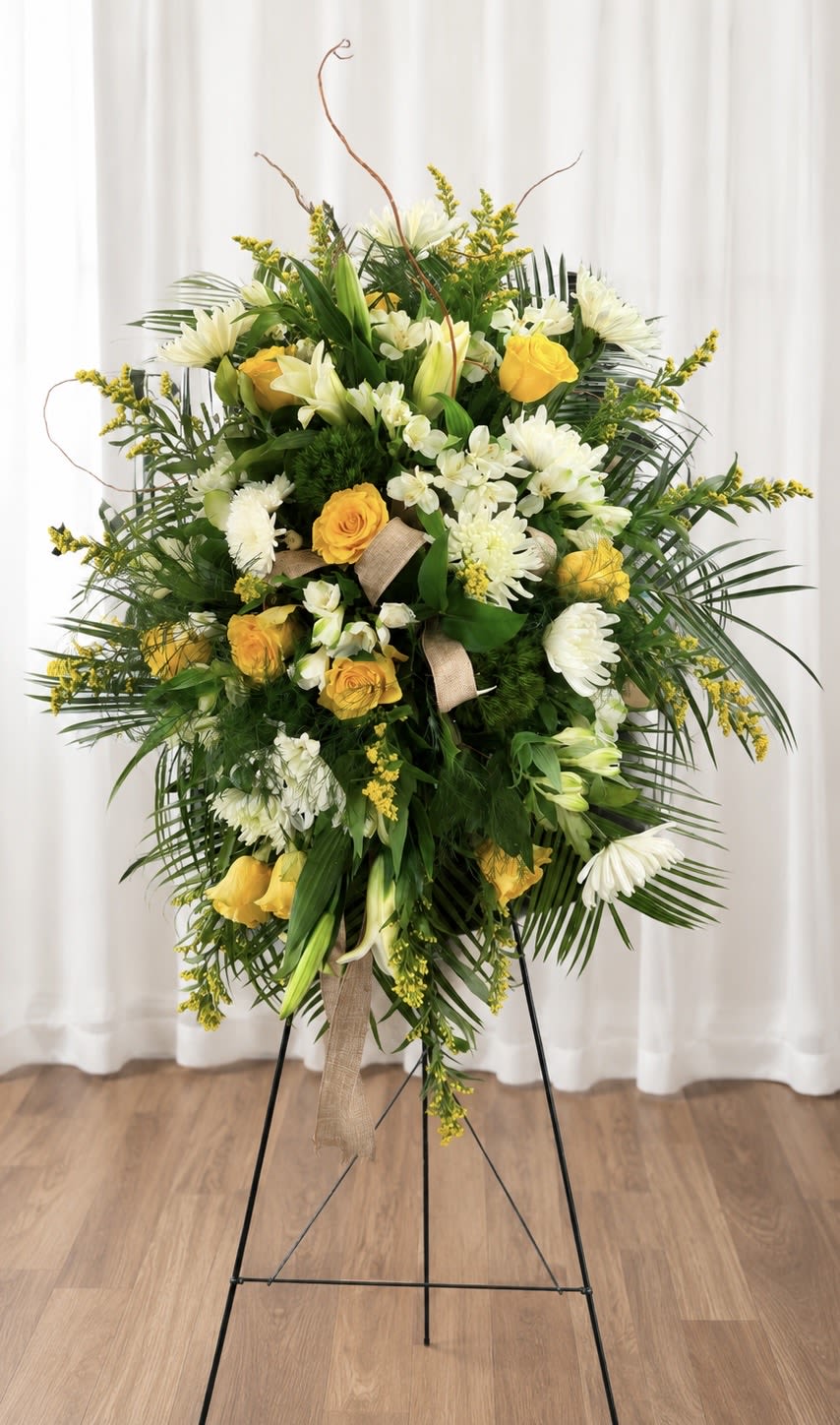 Golden Meadow Tribute - Soft golden roses and pure white lilies form a dignified standing spray designed to comfort and honor - a sympathy floral arrangement for memorial services and heartfelt remembrances. The vertical, layered composition on a black metal easel reads formal and ceremonial, the kind of tribute that helps the person you're thinking of feel held and remembered.  This large arrangement pairs yellow roses, white lilies, white chrysanthemums, alstroemeria, and delicate white accent blooms with abundant palm-like greenery and yellow filler flowers for depth and texture. A beige ribbon tied near the center adds a gentle, respectful touch; the full, upright silhouette creates a striking presence at a service or graveside.  Ideal when you want to express sympathy, offer condolences, or mark a memorial, this piece conveys warmth without overpowering the moment. Because it's mounted on a sturdy black easel, it arrives ready to display; if you need delivery timing or placement guidance for a service, we're happy to help coordinate.   If any design elements are of major importance to your order, please include them in the florist instructions at checkout or contact us to ensure availability.