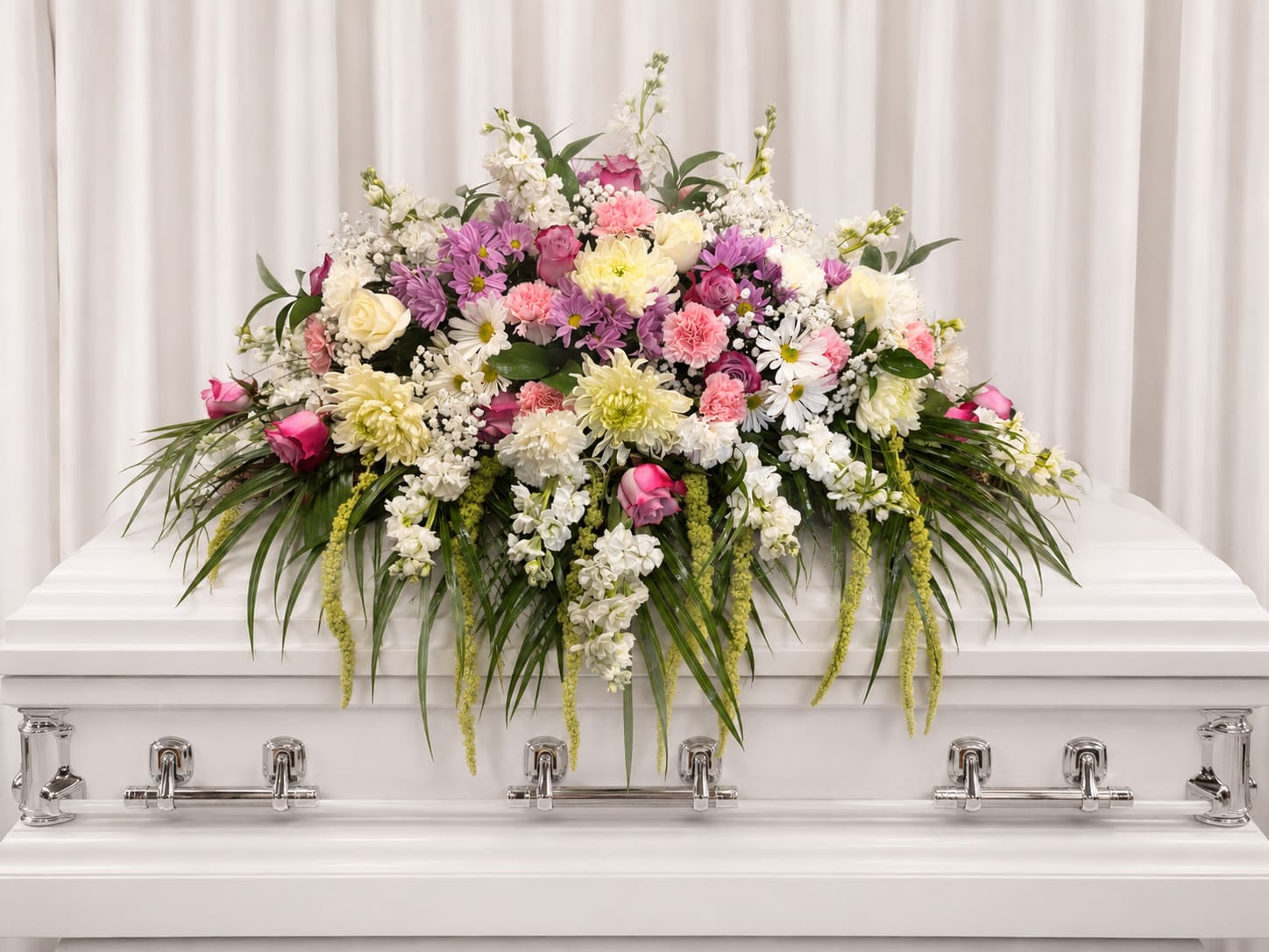 Beautiful faith - Soft white roses and blushing pink carnations form a serene, full altar piece designed for sacred moments-perfect as a wedding altar arrangement or a memorial flower for a chapel service. The composition reads reverent and celebratory at once, so the person you're honoring will feel surrounded by gentle color and thoughtful presence.  This large, low sympathy arrangement features a dense mix of white, pink, and purple blooms-roses, chrysanthemums, daisies, carnations, and complementary seasonal flowers-layered over long, arching greenery that cascades gracefully over the front edge. Placed on an altar table with a white daisy-like blossoms create a lush, textured sweep that fills the front of the altar without towering above it.  Ideal for a funeral, or commemorative service, this piece offers a peaceful, uplifted atmosphere that helps guests focus on the moment. Because it's designed to sit low and wide, it works well where visibility and approachability matter; please let us know altar dimensions or delivery/setup needs when ordering so we can ensure the perfect fit.  If any design elements are of major importance to your order, please include them in the florist instructions at checkout or contact us to ensure availability. 