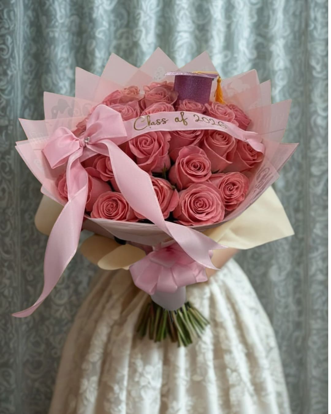 Graduation Glam Bouquet - Celebrate her big achievement with our Graduation Glam Bouquet, featuring 25 stunning light pink roses beautifully wrapped in elegant blush tones and finished with a satin ribbon. Topped with a charming graduation cap and personalized Class of 2026 detail, this bouquet is the perfect mix of elegance, sweetness, and celebration for your graduate’s special day.