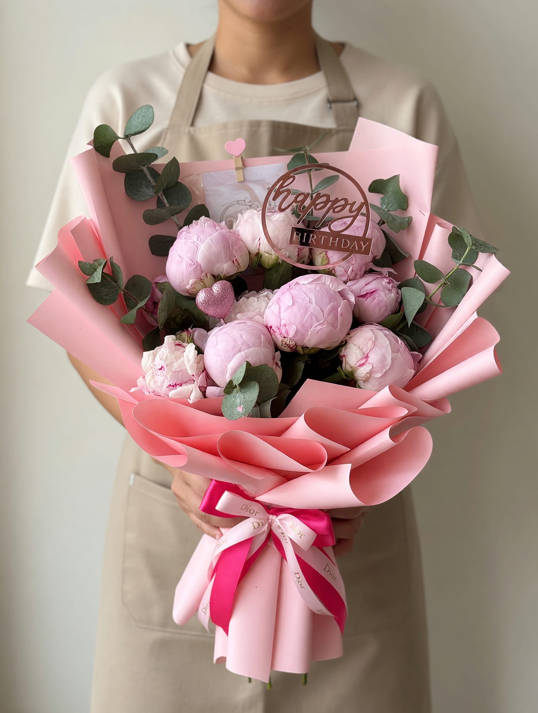 10 Peonies Bouquet  - Large blush-pink peonies take center stage in this birthday bouquet, creating a soft, romantic focal that feels both feminine and freshly joyful. As a birthday bouquet or cheerful everyday gift, the rounded spray of ten peonies-accented with a few crisp white blooms-sends a clear message of celebration and affection for the person you're honoring.  Muted eucalyptus branches weave cool green contrast through the lush pink heads, adding texture and a natural, airy silhouette. The stems are finished with layered pink wrapping paper, a decorative ribbon, a circular "happy birthday" topper and a small heart pick for a polished, personal presentation that arrives ready to gift.  Perfect for moms, sisters, close friends, or anyone who delights in pretty, fragrant flowers, this arrangement makes the recipient feel truly celebrated. Arrange on a bedside table or kitchen counter right away; peonies prefer fresh water and a cool spot away from direct sun to extend their vase life.