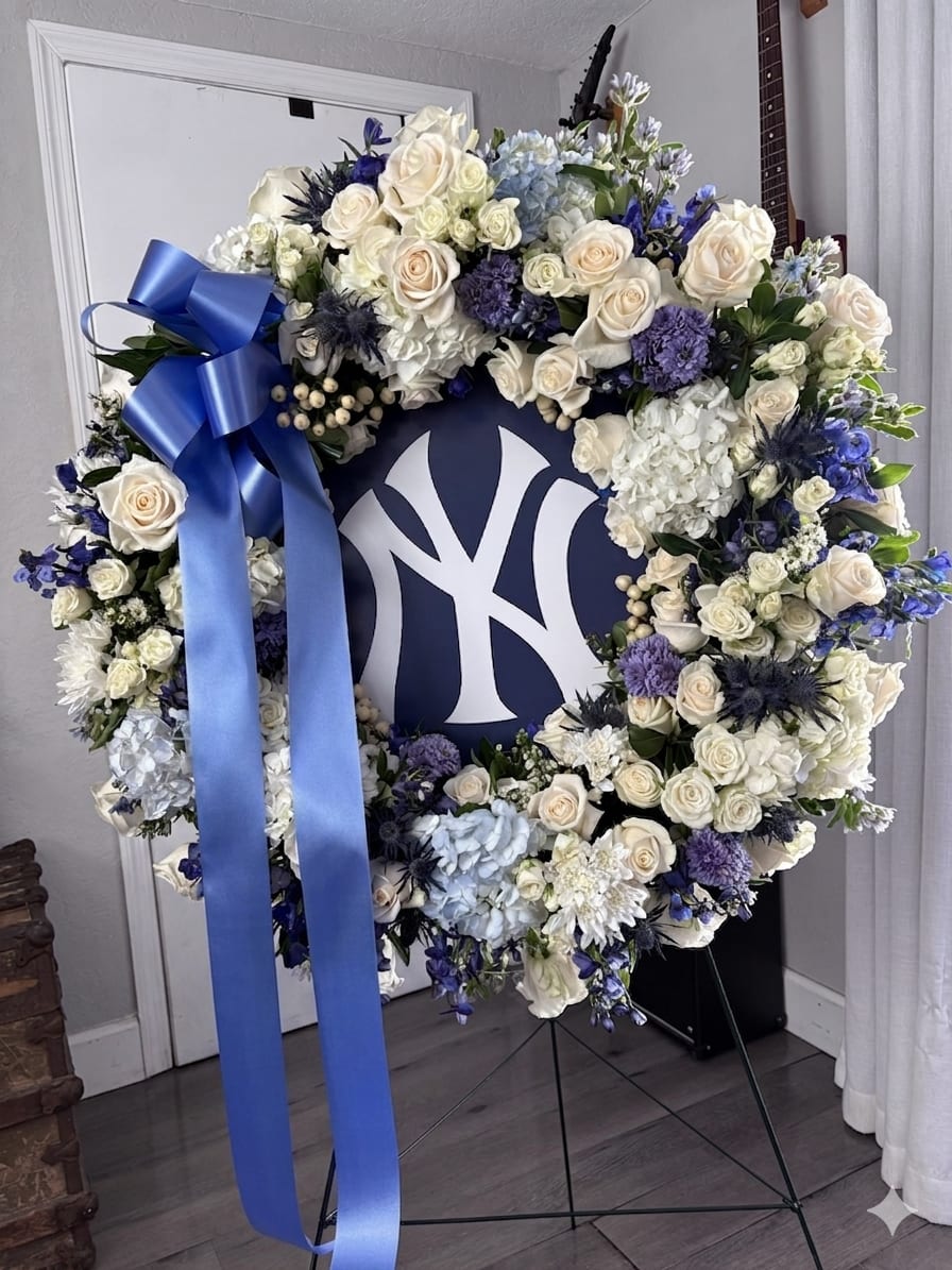 Customizable Funeral wreath  - A large  funeral wreath that honors a life and a fandom at once-this customizable funeral wreath pairs white roses, pale blue hydrangeas and purple accent blooms in a formal tribute wreath perfect for a memorial or celebration of life. Built around a navy-and-white team logo, this arrangement reads as both respectful and proudly personal-the kind of tribute that makes the person you're remembering feel seen for who they were.  The wreath is crafted on a sturdy frame with white roses, soft blue hydrangeas, and mixed chrysanthemums and other seasonal flowers woven for fullness and texture; a satin blue ribbon cascades down the left side for a polished finish. Shown on a wreath stand with a prominent sports logo sign at center, dimensions are generous to suit display at a service or graveside; everything on the wreath is customizable to match your chosen colors, flowers, ribbon style, or team sign.  Made to comfort family and friends, this wreath is ideal for a memorial service, funeral home display, or a special tribute at a game-day memorial. Tell us the colors, flowers, or logo you prefer and we'll personalize each detail-our team will advise on substitutions and sizing so the finished tribute arrives arranged, secured, and ready for display.