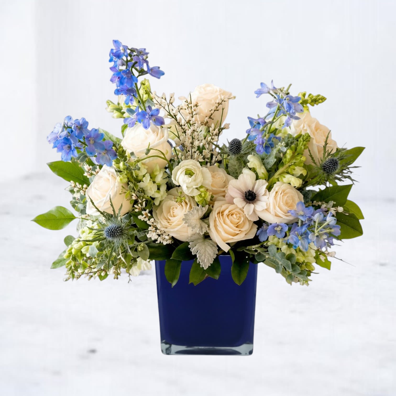 Sapphire - Sapphire is a refined blue and white floral arrangement designed with ivory roses, blue delphinium, soft seasonal blooms, textured thistle, and lush greenery in a striking cobalt blue vase. Inspired by coastal tones and airy garden florals, this elegant arrangement brings a fresh, calming presence to birthdays, sympathy gestures, anniversaries, thank-you gifts, or everyday flower delivery.  The rich sapphire cube paired with delicate blue florals creates a modern yet timeless design filled with movement, texture, and soft contrast. Designed in an organic garden style using premium seasonal flowers, Sapphire feels both sophisticated and effortlessly natural.  Same-day flower delivery available in Magnolia, The Woodlands, Montgomery, Conroe, Spring, Tomball, and surrounding Houston areas.
