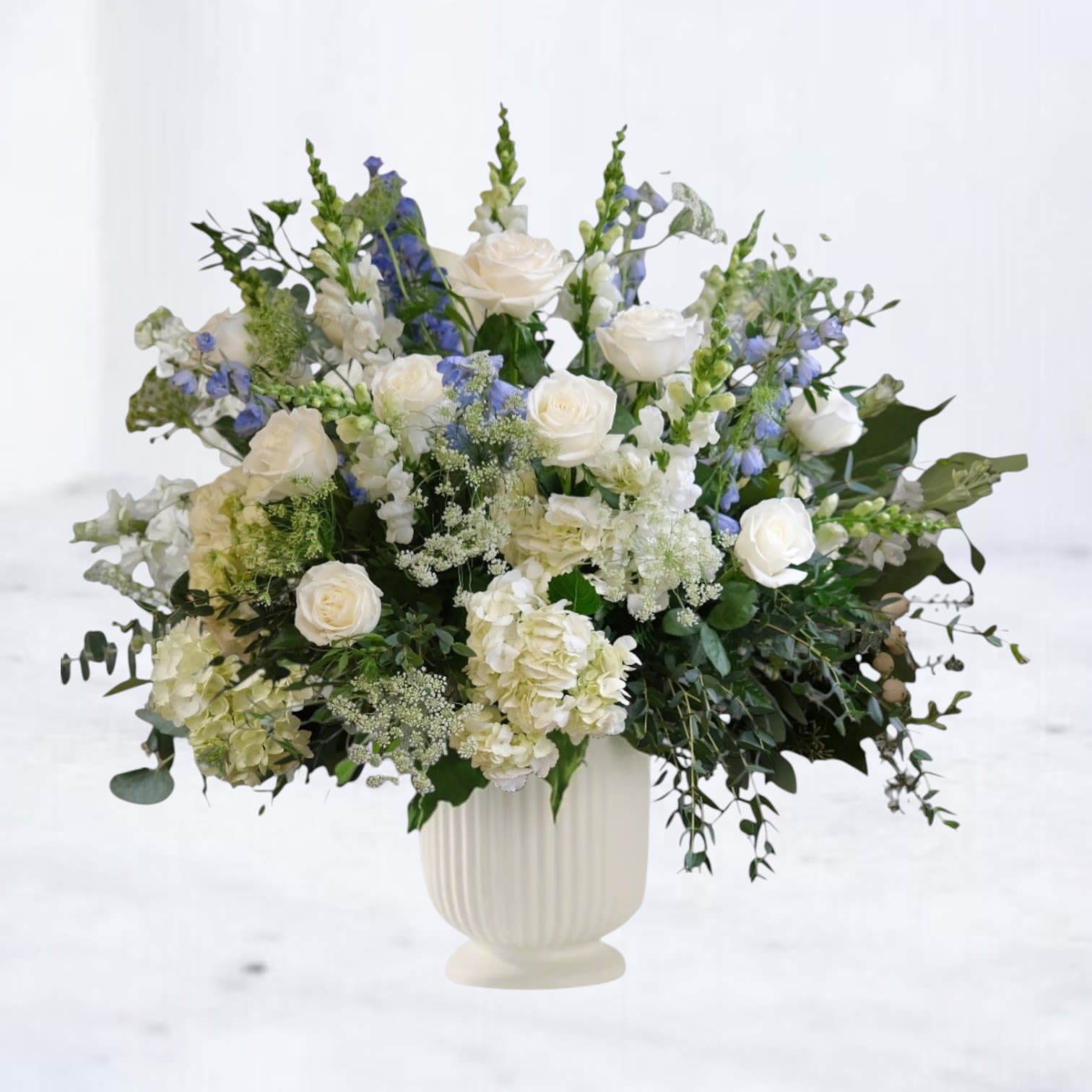 Eloria - Eloria is a serene funeral service arrangement designed to express remembrance, peace, and quiet grace. A harmonious blend of soft blues, gentle yellows, creamy whites, and layered greenery creates a composition that feels uplifting yet respectful — offering comfort without heaviness.  Designed in a classic urn vessel, Eloria is well suited for funeral services and memorials where families wish to honor a life with light, warmth, and calm presence.