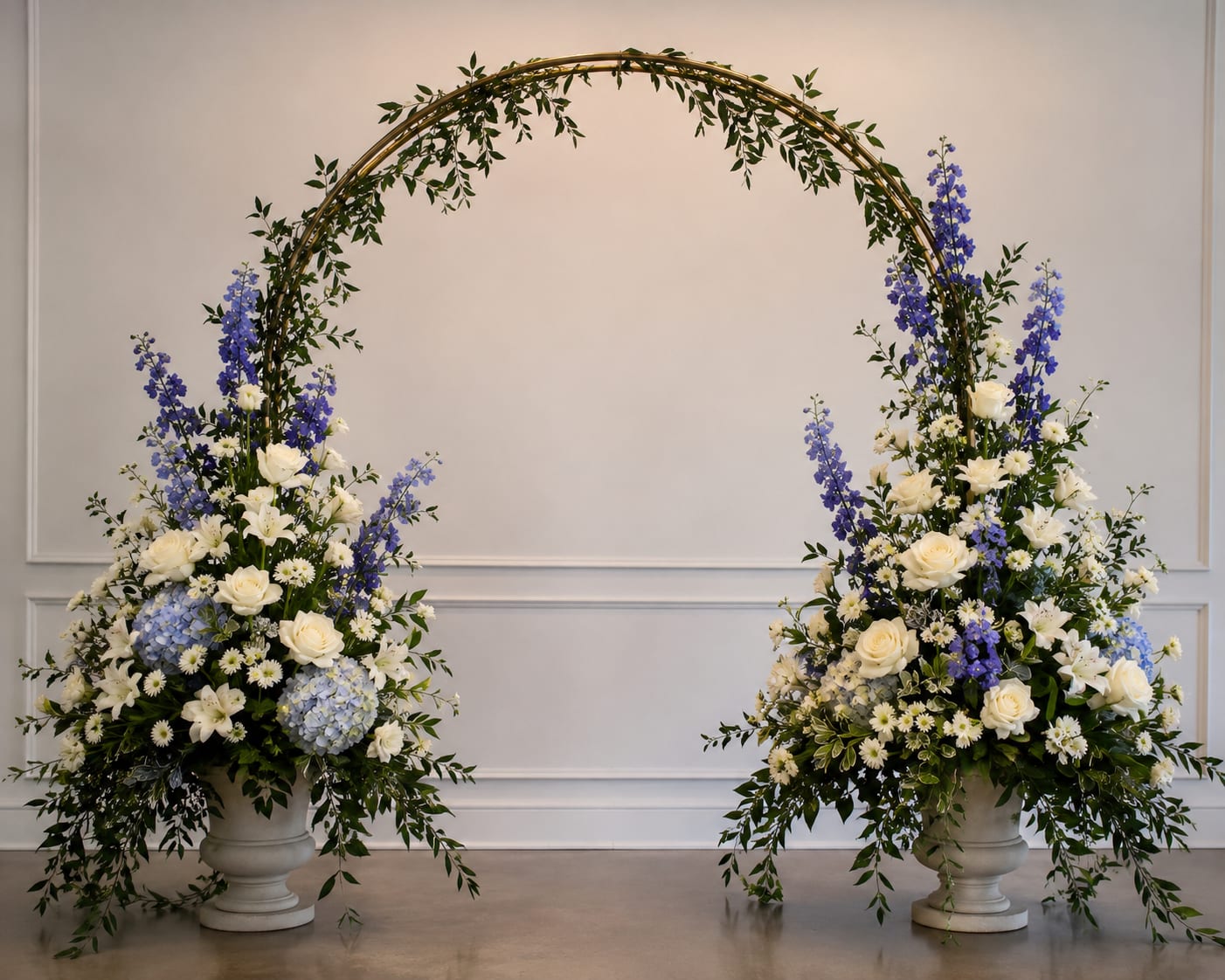 Honeymoon - A sweeping ceremony-ready display, Honeymoon pairs two tall floral arrangements on white pedestal urns linked by a graceful curved metal arch-perfect as a wedding backdrop or formal event focal point. The cool-toned palette of white roses and lilies with blue hydrangeas, sky-blue delphinium, and small daisy-like blooms reads as refined and romantic; the recipient will feel seen by an arrangement that suggests both celebration and quiet elegance.  Each pedestal arrangement is tall and airy, built with asymmetrical groupings that let the arched greenery breathe while showcasing focal roses and lilies against clusters of hydrangea and vertical delphinium. Textures move from the lush, cloud-like hydrangeas to the sleek petals of lilies and the delicate centers of the daisy-like flowers, all tied together with verdant greenery woven along the metal arch for continuity   Honeymoon delivers a refined backdrop that photographs beautifully and makes guests feel the moment. Order to reserve the pedestals and arch for your date; because these are tall, ceremony-style pieces, they're intended as statement décor rather than a tabletop centerpiece-ask us about delivery and installation options to ensure perfect placement.   If any design elements are of major importance to your order, please include them in the florist instructions at checkout or contact us to ensure availability.