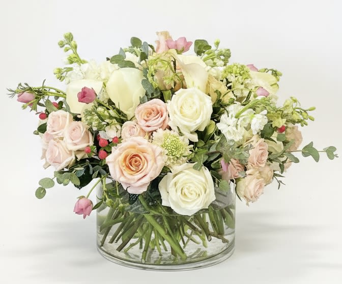 Elsa - Pale blush and peach roses mingle with soft white lisianthus and airy stock in a garden-style bouquet made to feel like a quiet, romantic moment. This anniversary bouquet (also perfect for weddings or sympathy gifting) delivers a gentle, comforting presence-the person you're thinking of will feel seen and softly celebrated by its tender palette of white, pink, peach, and touches of green.  The arrangement is full and rounded but deliberately loose, with stems and small filler blooms extending outward for a soft, abundant silhouette. Roses, lisianthus, and stock create layers of petal texture and a subtle fragrance, all presented in a clear glass vase that keeps the composition light and shows the gathered stems inside for a fresh, natural look.  Choose this arrangement when you want to mark love, support, or remembrance with understated elegance-the kind of bouquet that brings calm and warmth into a room. The vase is included and sized to support the full, garden-like shape; to keep blooms looking their best, refresh the water every two days and trim stems at an angle if you want to extend their vase life.
