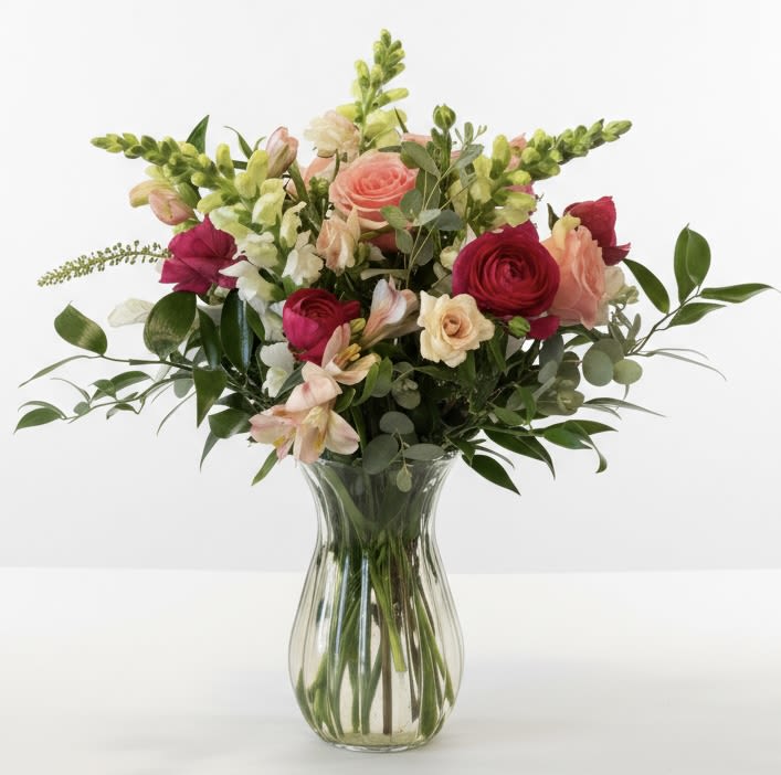 Stella - Soft pink roses and peach blooms mingle with creamy white alstroemeria and upright snapdragons in a garden-style bouquet that feels both romantic and effortlessly joyful-perfect as a birthday flower arrangement or for an anniversary that calls for gentle, heartfelt color. The clear glass vase lets the stems show through, giving the mix a fresh, natural look that reads as "just because" sweetness or a tender Valentine's Day surprise without feeling fussy.  Rounded rose heads sit beside clustered alstroemeria and tall, airy snapdragon spikes to create a full yet breathable shape. The palette of bright pink accents, pale blush and cream tones, plus fresh green foliage, keeps the arrangement lively and versatile. Sized to sit beautifully on a dining table or bedside, the bouquet arrives arranged in a simple clear glass vase for immediate display.  Give this to someone you want to make feel cherished-she'll feel noticed, loved, and celebrated whether it's for a birthday, an anniversary, or a spontaneous moment of romance. For best freshness, keep the vase topped up with water and trim stems every few days to prolong bloom life.