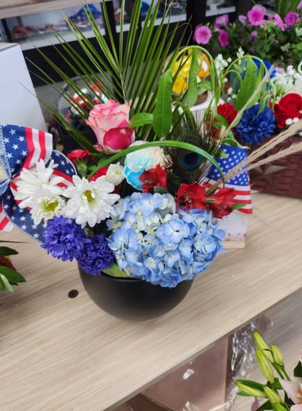 Red, White &amp; Blue Floral Arrangement - Vivid clusters of blue hydrangea and bold red blooms make a striking first impression in this patriotic flower arrangement - perfect as a Fourth of July centerpiece or a summer celebration bouquet. Designed as a festive flower arrangement for Independence Day, Memorial Day, or any backyard barbecue, the American flag ribbon and bright color story say "celebrate" the moment your person walks in.  This design is arranged in a rounded black vase and packed with blue hydrangeas, white daisy-like blooms, red flowers, pink accents, and a few yellow highlights for extra cheer. Tall palm-like fronds and looping green leaves lift the composition for a full, airy silhouette that reads as both casual and carefully curated. The flag ribbons on either side add a classic, seasonal touch that ties the whole palette together.  Give this to the neighbor who hosts every summer gathering, a veteran you want to honor, or anyone who loves bold, patriotic color - they'll feel genuinely celebrated. Vase-ready and arranged for immediate display; keep in cool water away from direct sun to extend freshness.