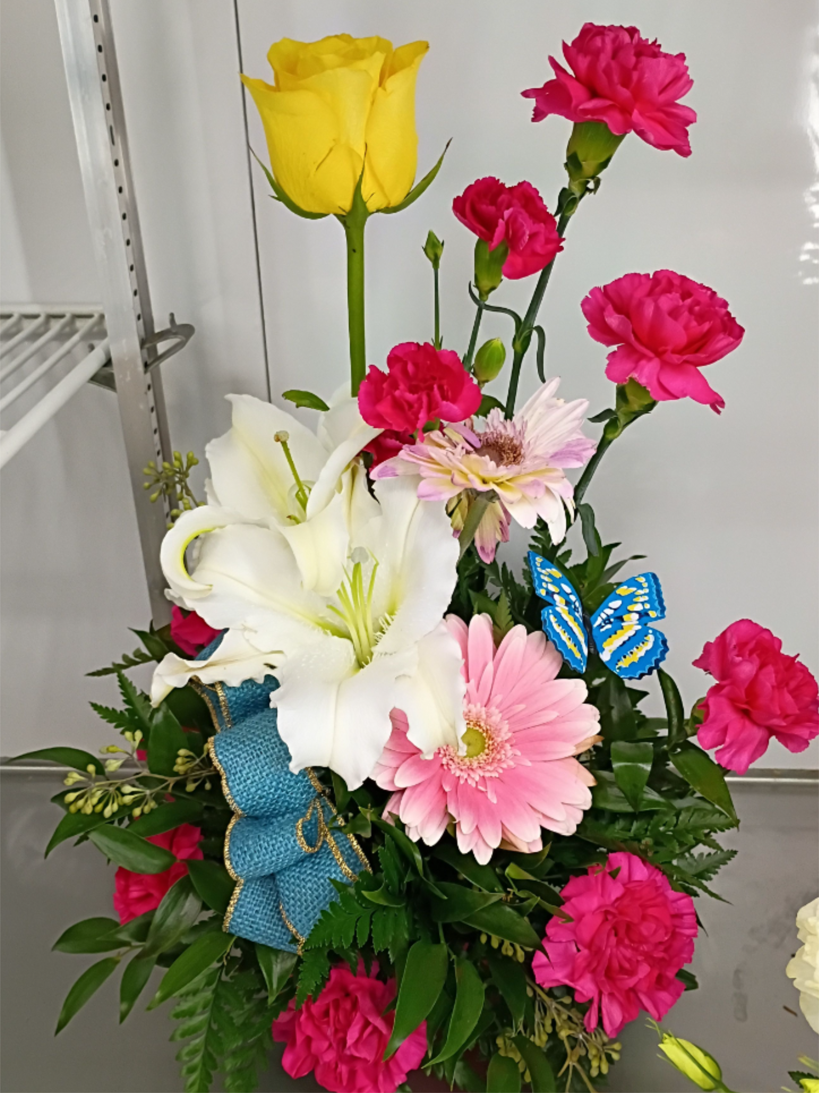 Short gift arrangement - A tall yellow rose crowns this short gift arrangement, surrounded by fragrant white lilies, bright pink carnations, and soft pale-pink gerbera daisies for a cheerful birthday bouquet or congratulatory gift. The mix of white and vivid pink creates a lively contrast that reads celebratory and hopeful-perfect for making the person you're thinking of feel seen and celebrated.  Flowers are arranged upright in a dense, layered design so the lilies and rose create a warm focal height while the carnations and gerbera add texture and fullness at mid and lower levels. A textured blue ribbon with coordinating blue burlap ribbon is tied at the base and a small decorative butterfly pick adds a playful finish; the palette includes touches of blue along with pink, white, yellow, and mixed color accents.  Ideal for birthdays, congratulations, or a bright just-because surprise, this arrangement arrives ready to display and brings a polished, gift-like presentation. Care: keep blooms in fresh water, trim stems every few days, and place away from direct heat to prolong vase life.