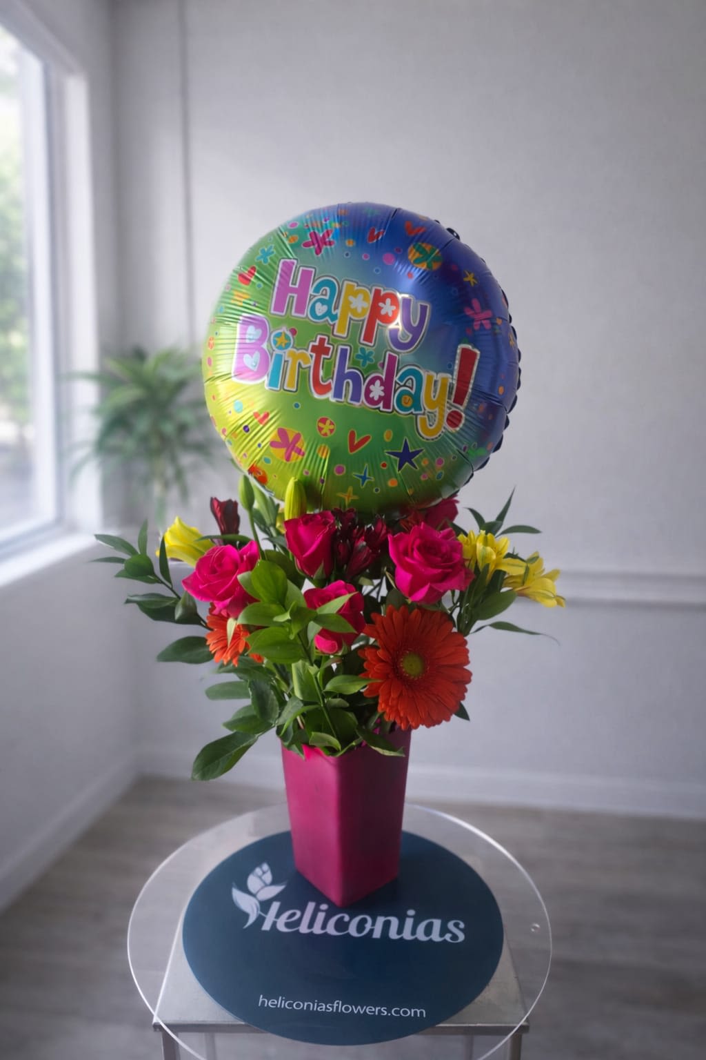 Fuchsia Birthday Bloom - A tall pink vase holds a compact, celebratory bouquet topped with a bright birthday foil balloon-perfect as a birthday arrangement or for any joyful occasion. The festive mix of hot pink roses, a red gerbera daisy, yellow blooms, and orange accents reads playful and gift-like; the balloon adds height and makes the display feel like a party-ready presentation that will make the person you're celebrating feel truly special.  This arrangement pairs roses, gerbera daisies, and alstroemeria in a lively palette of pink, red, yellow, and warm mixed tones, arranged tightly for a bold visual impact. The pink vase brings extra color continuity and keeps the design upright and elegant, while the foil balloon gives a fun vertical focal point. Ideal for birthdays, congratulations, or simply to brighten someone's day-order with standard sizing as shown; vase and balloon are included, and simple water refreshes will keep the blooms looking fresh for several days.