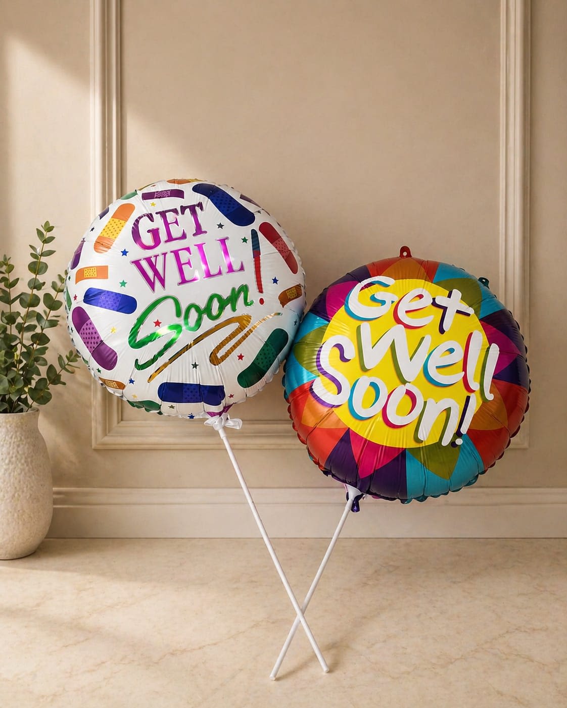 Get Well Soon Balloons - Cheerful paired foil balloons that say "Get Well Soon!" make an upbeat, non-floral gift perfect for recovery visits or to brighten someone feeling under the weather. This balloon arrangement is ideal for a hospital room, home recovery, or as a small encouragement pick-me-up-the colorful messages and playful bandage graphics immediately signal care and good wishes to the person you're thinking of.  Two round foil balloons are included: one with a white background, multicolored bandage motifs and bold purple-and-green lettering, the other featuring a rainbow border and sunny yellow center. Both come mounted on balloon sticks for easy display without helium, creating a tidy, portable presentation that nurses or family can place bedside or on a table. Order this as a simple, thoughtful way to let someone know you're rooting for their recovery.