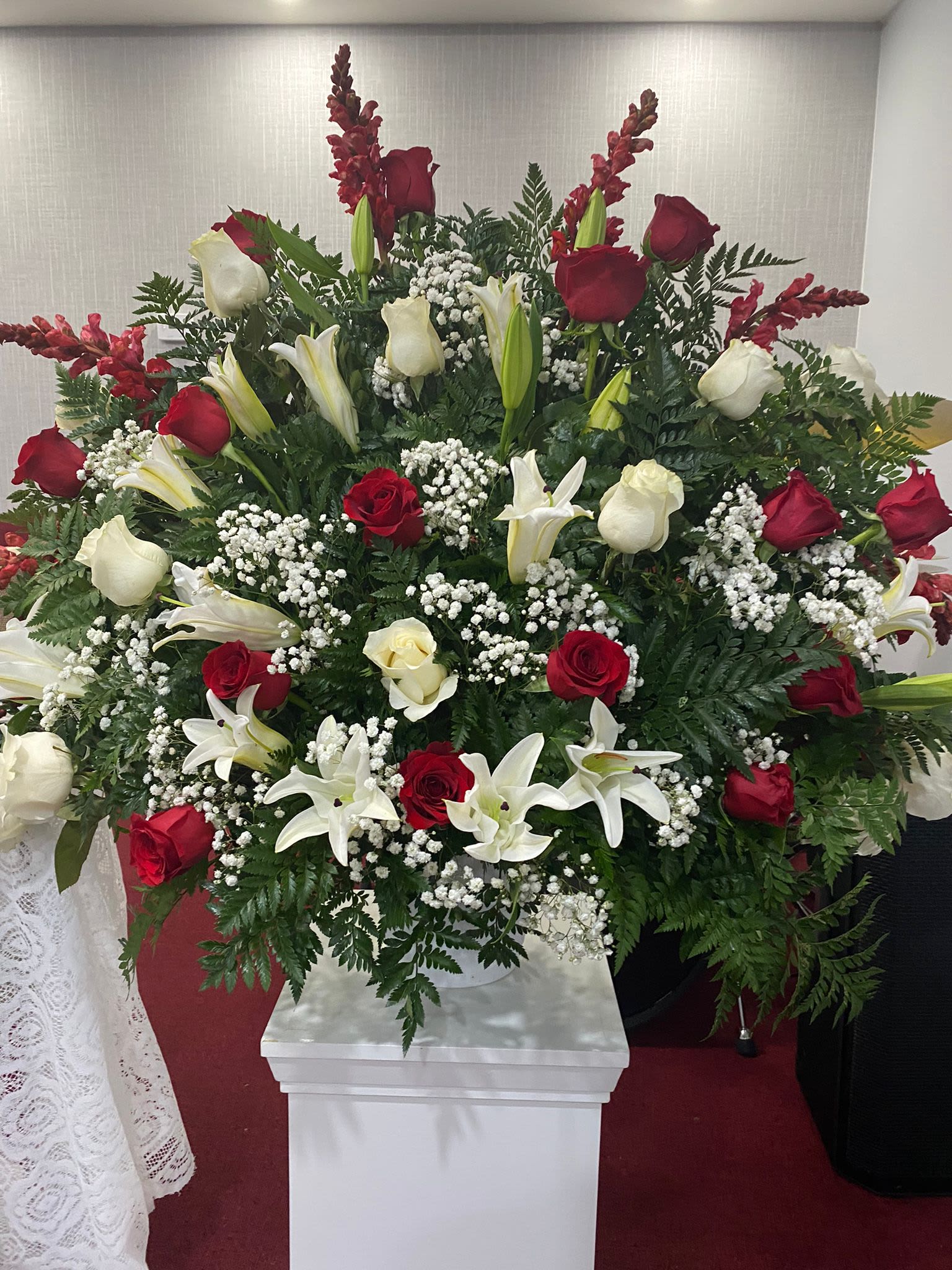 Arrangement for church ceremonies and special events - A formal, large-scale spray designed for church ceremonies and special events, this red, white, and green arrangement makes a dignified statement for memorial services or solemn celebrations. Built on a white pedestal stand, the composition centers on red and white roses paired with white lilies to create a classic, ceremonial look that reads clearly from the front pews - the kind of floral tribute that helps the person you're honoring feel seen and remembered.  Dense, layered blooms form a broad, rounded base accented by fern-like greenery and clusters of baby's breath for soft texture. Tall red accent spikes rise above the massed flowers to give height and vertical presence; the lilies add light-reflecting white surfaces while the roses provide rich color and shape. This arrangement's scale and traditional styling suit formal settings and will coordinate with sanctuary decor without overwhelming the space.  For services or commemorative events, this spray offers a respectful, enduring focal point that conveys love and remembrance. Delivered on a sturdy white pedestal stand, it arrives ready for placement; please note sizing and delivery details at checkout to ensure timely setup for your ceremony.