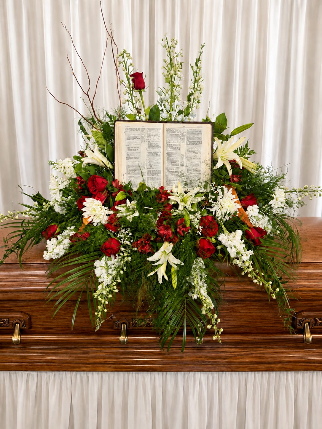 Promise Land - A large, reverent casket spray built around an open book creates an immediate focal point that speaks of faith and remembrance - ideal as a funeral arrangement or memorial tribute. Rich red roses and pure white lilies form a striking red-and-white contrast set against abundant greenery, giving the person you're honoring a dignified, comforting presence at the service.  This broad, cascading design includes white spray flowers and fuller white blooms woven with trailing foliage that drapes gracefully over the front of the casket. Tall branching accents rise behind the open book for height and profile, while the mix of roses, lilies, chrysanthemums and other white florals adds texture and depth to the composition. The wooden casket elements are complemented by the keepsake book centerpiece for a lasting symbol of faith and memory.  Choose this arrangement to convey love, respect, and consolation to family and friends during a funeral or memorial service - it reads as a solemn, hope-filled tribute to a cherished life. If specific blooms or placement details matter to you, please note them in the florist instructions at checkout or contact us to confirm availability.