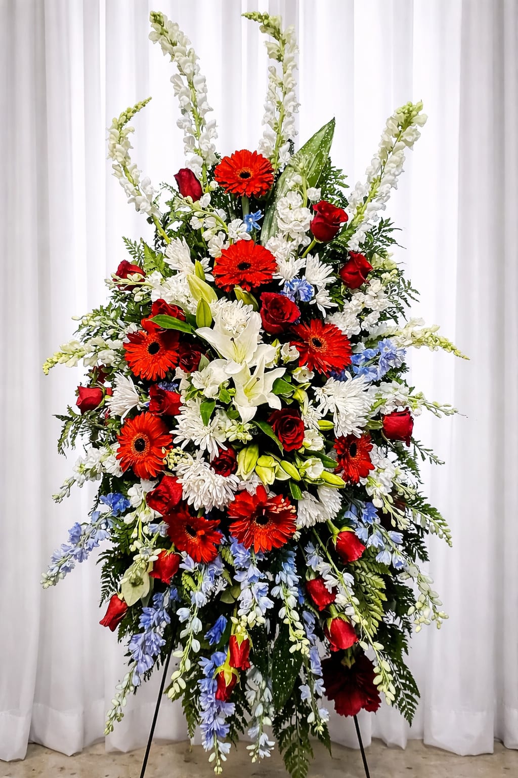 Glory - A bold, patriotic standing spray designed for a memorial or funeral, Glory presents a dramatic vertical silhouette that honors a life with dignity. The large standing easel frames bright red gerbera daisies and red roses alongside pure white lilies and daisy-like blooms, with pale blue accents woven through to create a red, white, and blue tribute that reads both ceremonial and heartfelt.  Tall white snapdragon spikes give stately height while a fuller, looser lower cascade softens the composition for a balanced, formal look. The mix of gerbera daisies, roses, lilies, daisies and other seasonal blooms delivers varied textures-from the lily's fragrant, open petals to the structured snapdragon spikes-so the arrangement reads clearly from a distance at a service or graveside.  Designed to stand prominently on an easel, Glory is the kind of sympathy tribute that helps the person you're remembering feel honored and seen. For ordering and sizing details or to customize colors or specific flowers, please contact us so we can make sure the spray suits the service and location.