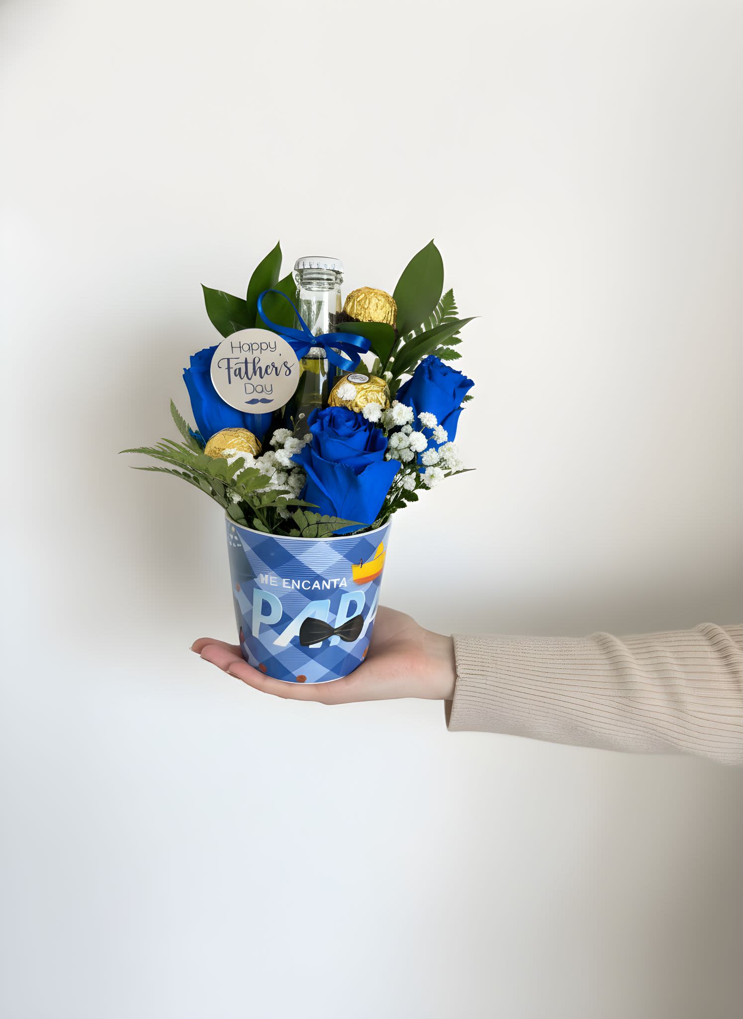 Dad Cup  - Vivid blue roses clustered with white filler blooms make this Father's Day flower arrangement a bold, celebratory gift that reads both festive and personal. Designed as a compact gift arrangement for Father's Day, birthdays, or a thank-you, the vivid blue-and-gold palette and round "Happy Father's Day" tag send a clear message of appreciation-perfect for the dad, stepdad, grandpa, or father-figure you want to honor.  The arrangement sits in a blue patterned paper cup wrapped with a bright blue ribbon and a round Father's Day tag, with glossy green foliage framing the flowers for contrast. Gold-wrapped Ferrero Rocher chocolate truffles are nestled around a central glass bottle for an extra touch of indulgence; the mix of roses, small white fillers, and chocolate makes it both attractive and tasty. Its compact size is easy to carry and present by hand, making it a cheerful, polished choice when you want the recipient to feel seen and celebrated.