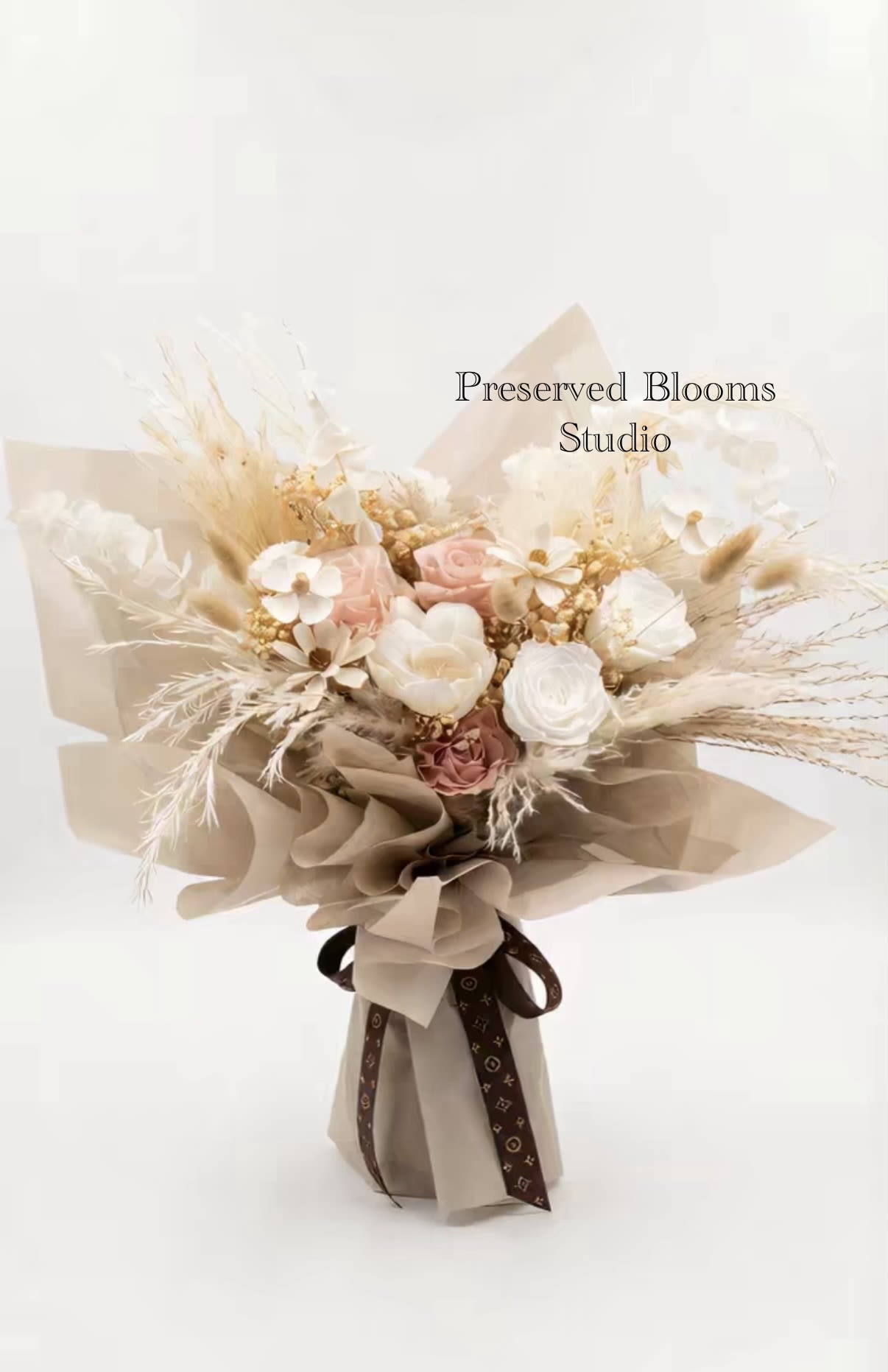 Bouquet of preserved white and blush flowers with dried grasses wrapped in tan paper and tied with a brown ribbon
