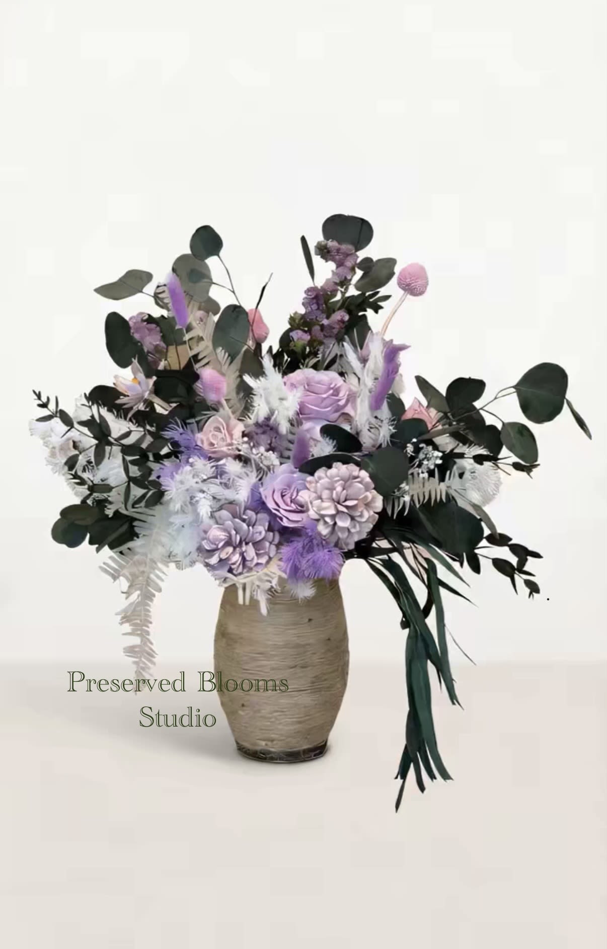 Tall arrangement of lavender and white preserved flowers with dark greenery in a textured beige vase