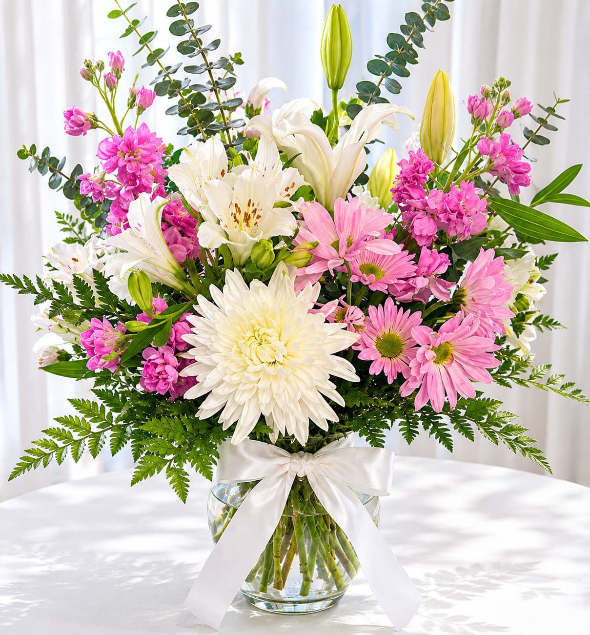 Majestic pink - Bright pink and soft white blooms make an immediate, celebratory impression - the perfect birthday bouquet or spring gift to make someone feel truly cherished. This upright flower arrangement pairs elegant white lilies with large white chrysanthemums and cheerful pink daisy-like blooms to create a feminine, joyful display that reads as both festive and refined.  Arranged in a clear glass vase and finished with a white satin ribbon, the composition is full-bodied with airy eucalyptus and fern-like foliage adding height, texture, and a fresh green contrast. The mix of lilies, chrysanthemums, daisies, and assorted filler flowers gives pleasing variety in shape and size while staying harmoniously pink, white, and green.  Send this to mark birthdays, congratulations, or any moment when you want the recipient to feel celebrated and seen; its bright palette suits springtime gifting but reads as appropriate year-round. The vase arrangement arrives ready to display - simply add fresh water and trim stems occasionally to keep blooms looking their best.   If any design elements are of major importance to your order, please include them in the florist instructions at checkout or contact us to ensure availability.